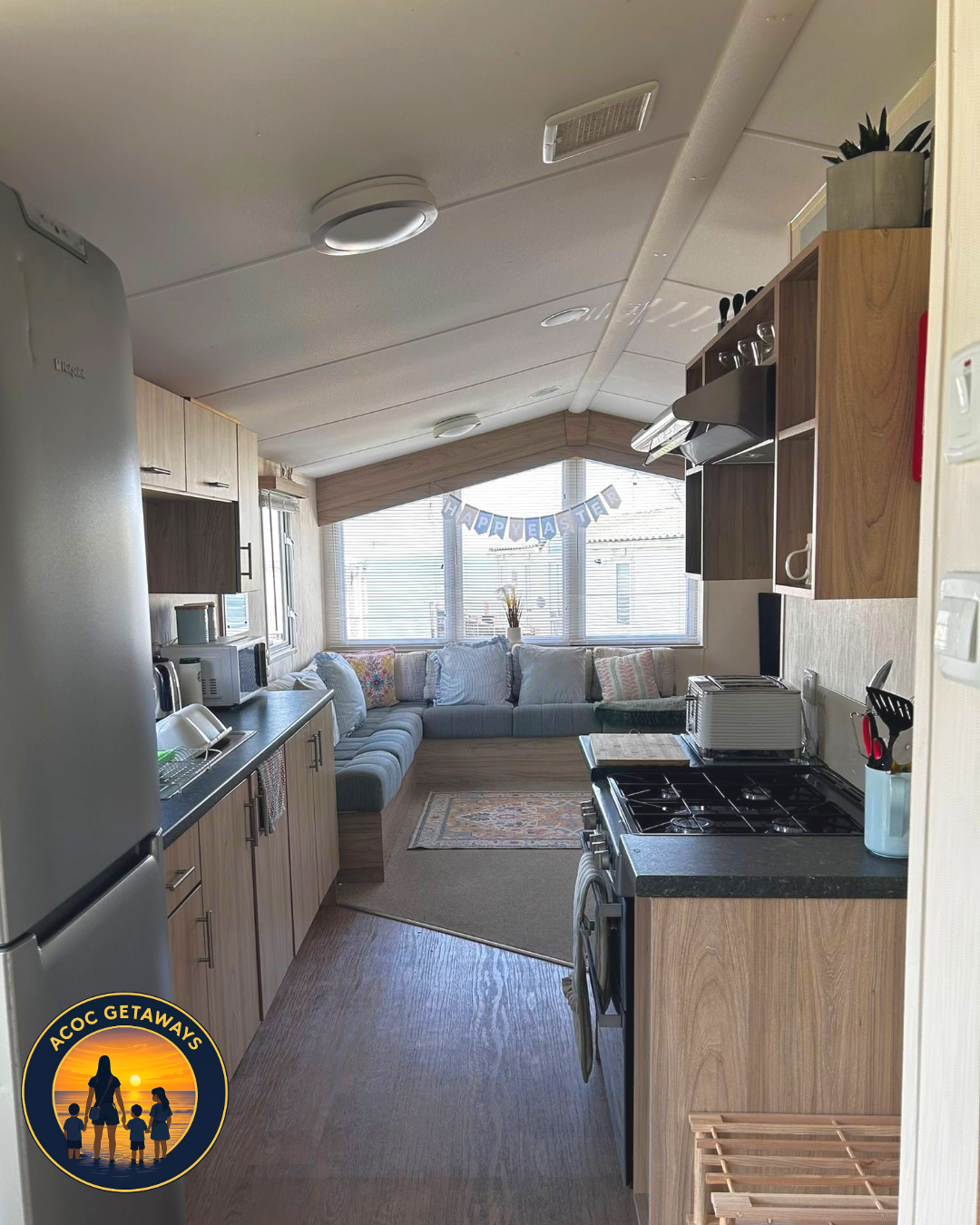Interior of a cozy camper with a kitchen area on the right and a sitting area with a large window at the back, decorated for Easter with a 'Happy Easter' banner.