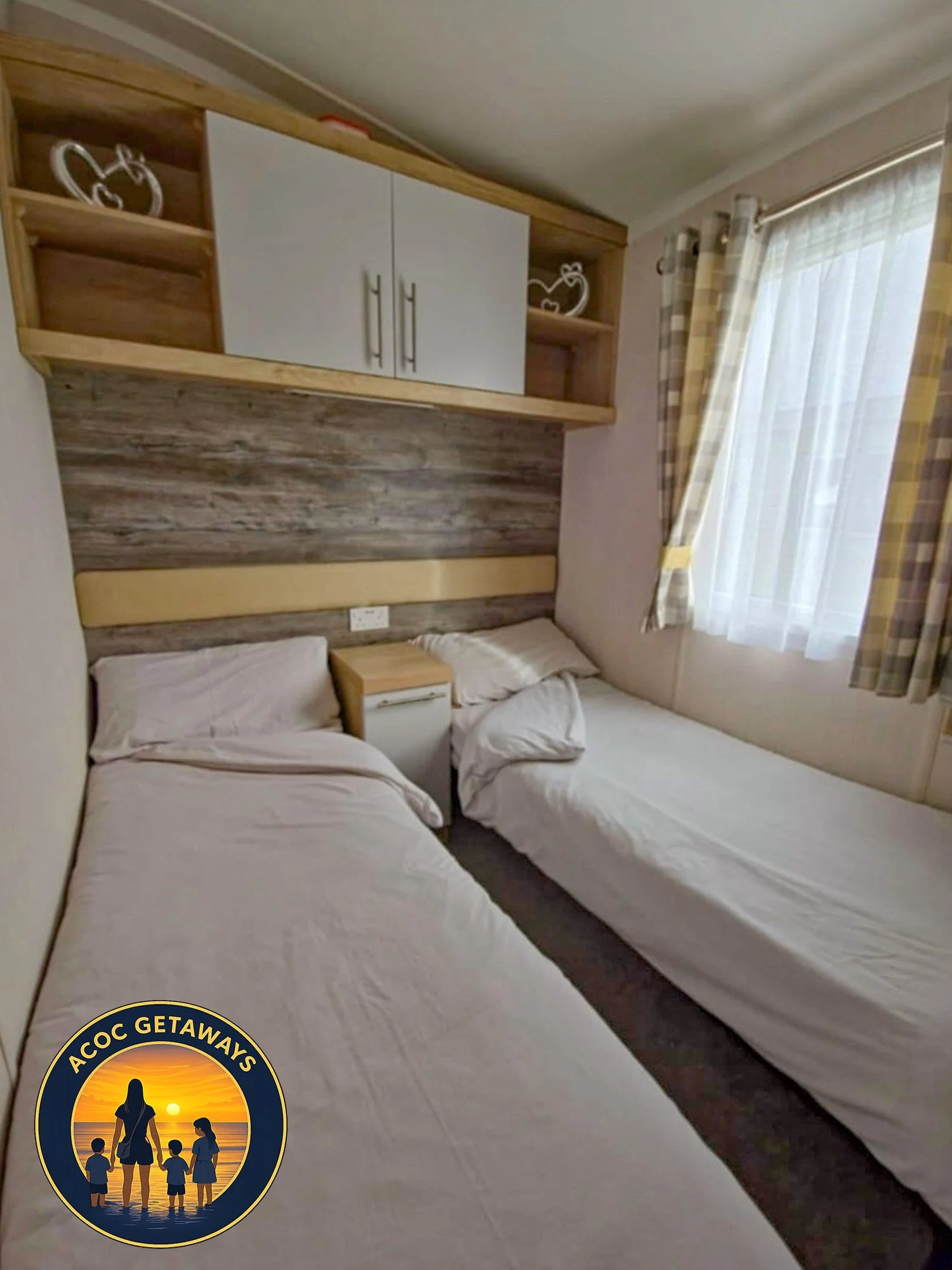 Hotel room with two twin beds, wooden headboard, nightstand, and window with checkered curtains.