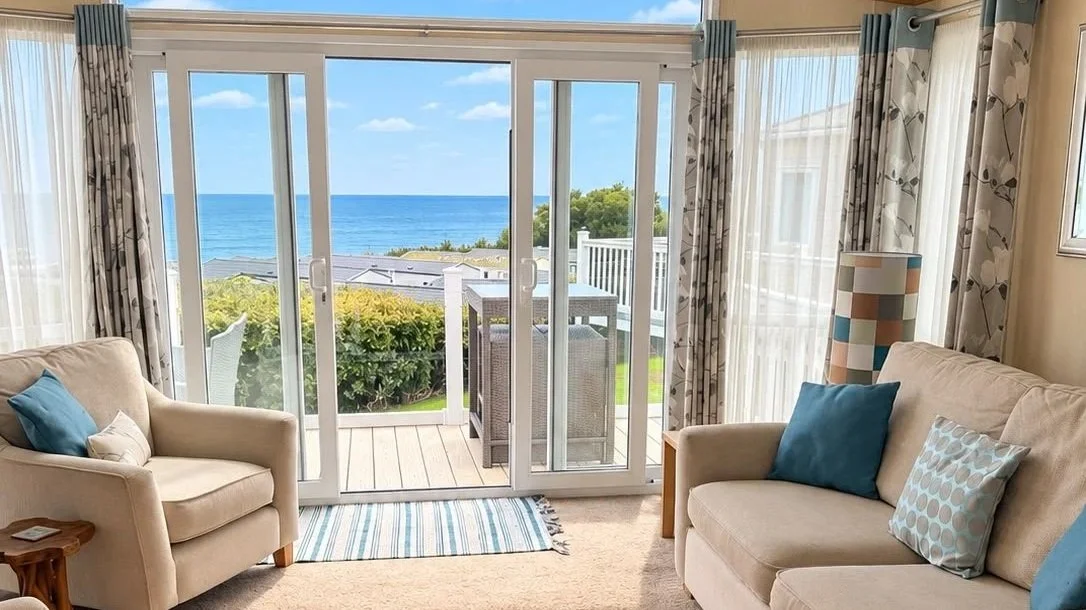 Living room with sliding glass doors leading to a balcony overlooking the ocean, with two beige sofas and blue pillows, patterned curtains, and a striped rug.