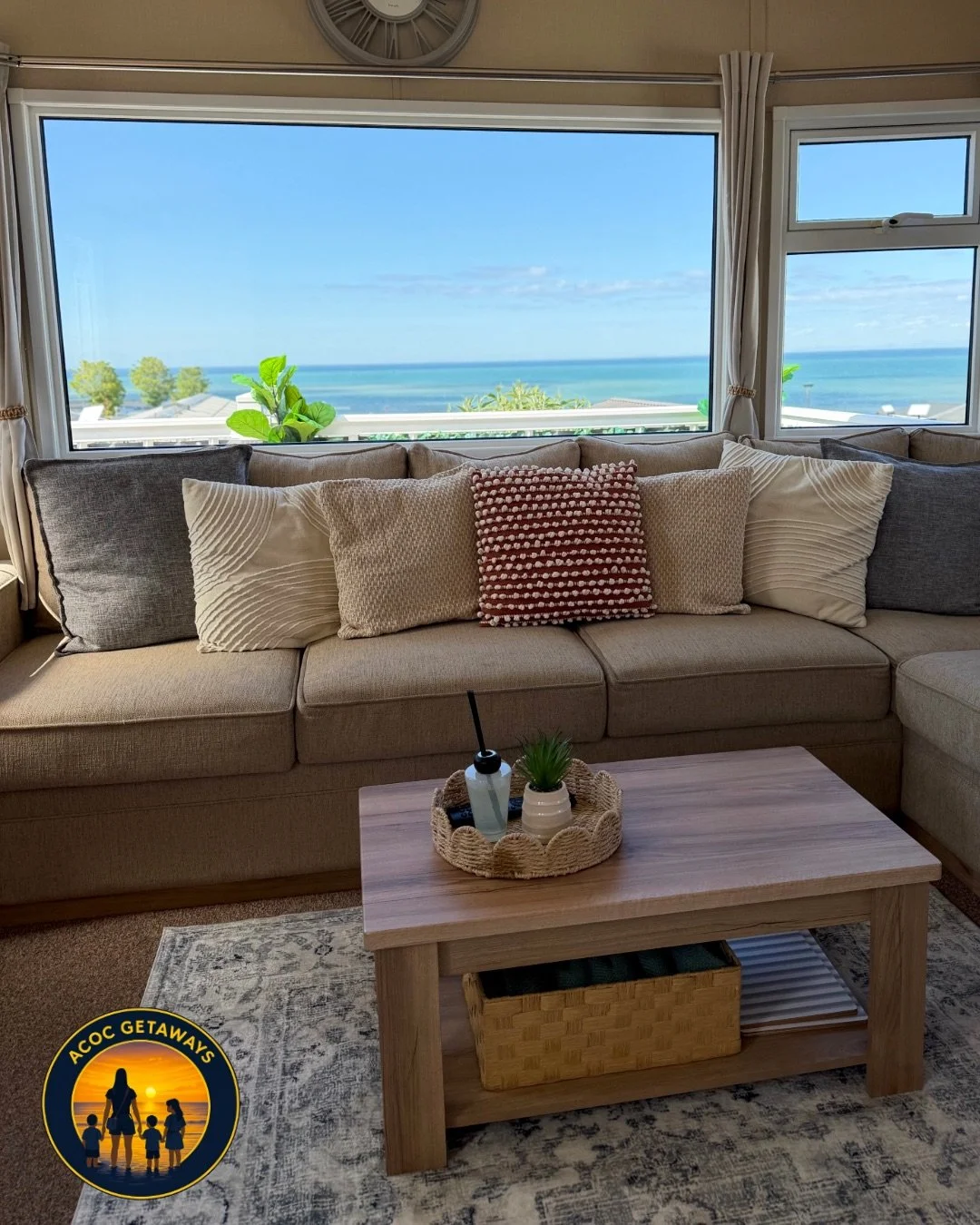 Living room with a beige sofa, decorative pillows, wooden coffee table with a small plant and tray, large windows showing ocean and sky view.