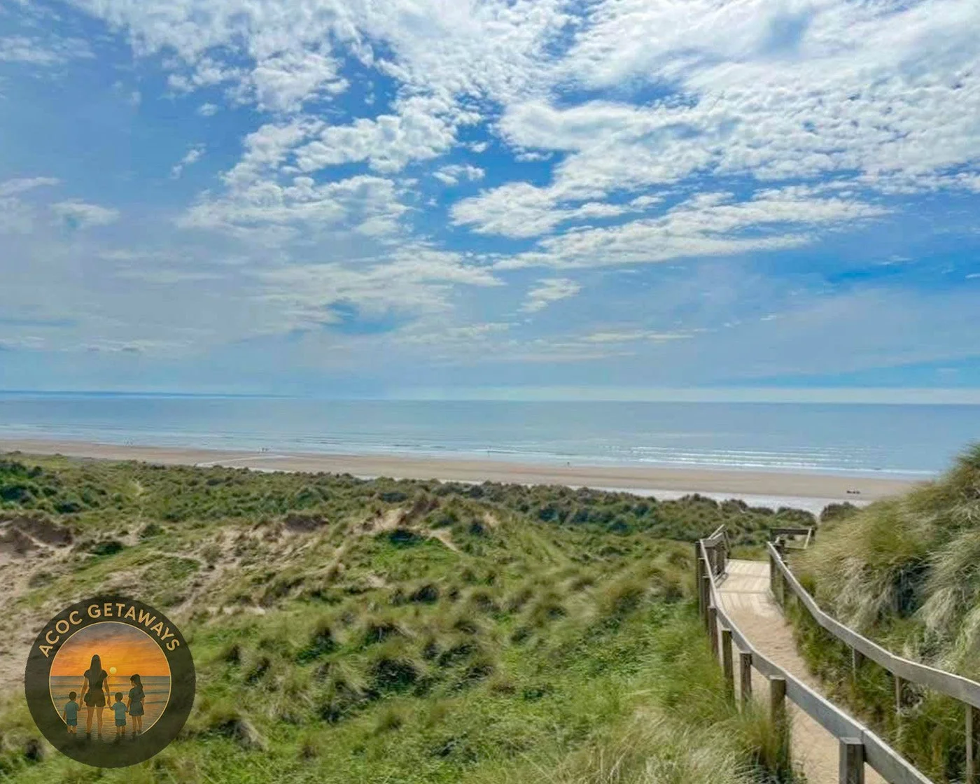 A pathway leading through grassy dunes towards a beach with calm ocean waters and a partly cloudy sky.