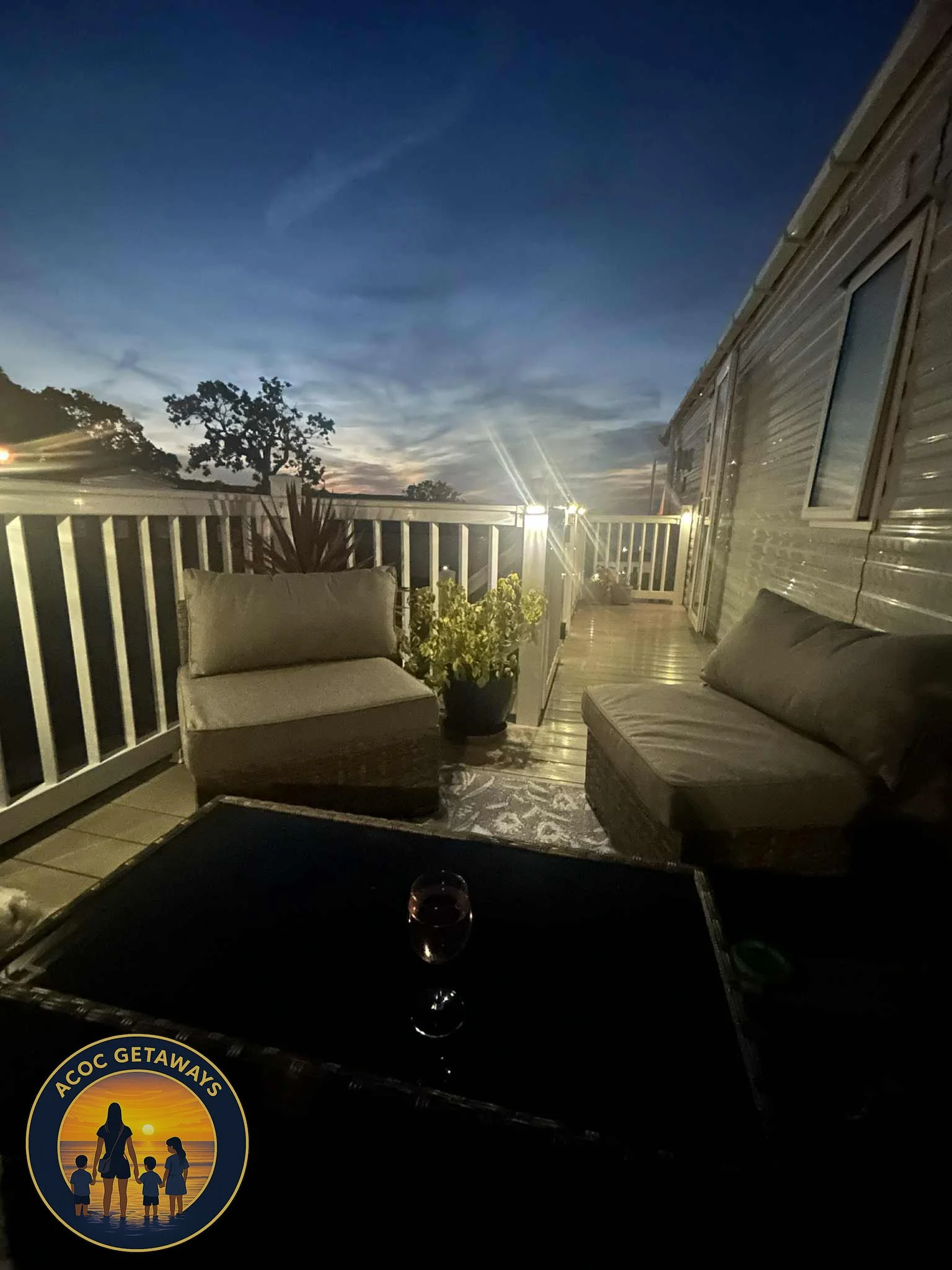 A balcony at dusk with outdoor seating, including a cushioned chair and a loveseat, a potted plant, and a rug. There are lights along the railing and a glass of wine on a table. The sky shows a sunset with blue and orange hues.