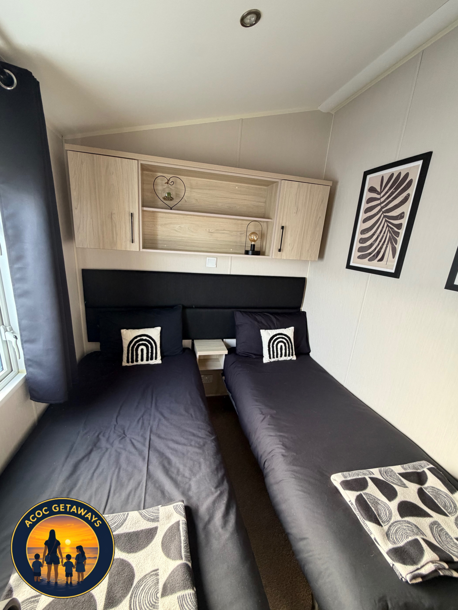 A room with two black beds, patterned pillows, a shelf with decorative items, framed artwork on the wall, and a small window with black curtains.