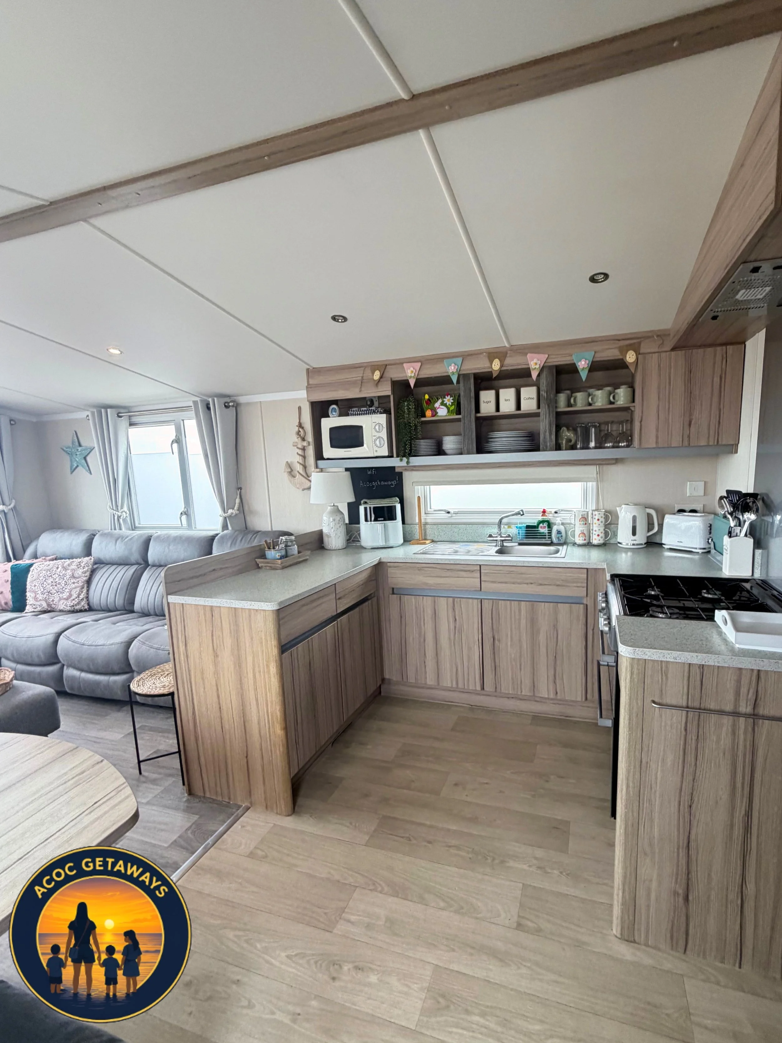 A cozy kitchen with wooden cabinets, a white countertop, and various kitchen appliances, located near a living area with a light gray sofa and decorative pillows, and a window with curtains and star-shaped ornament.