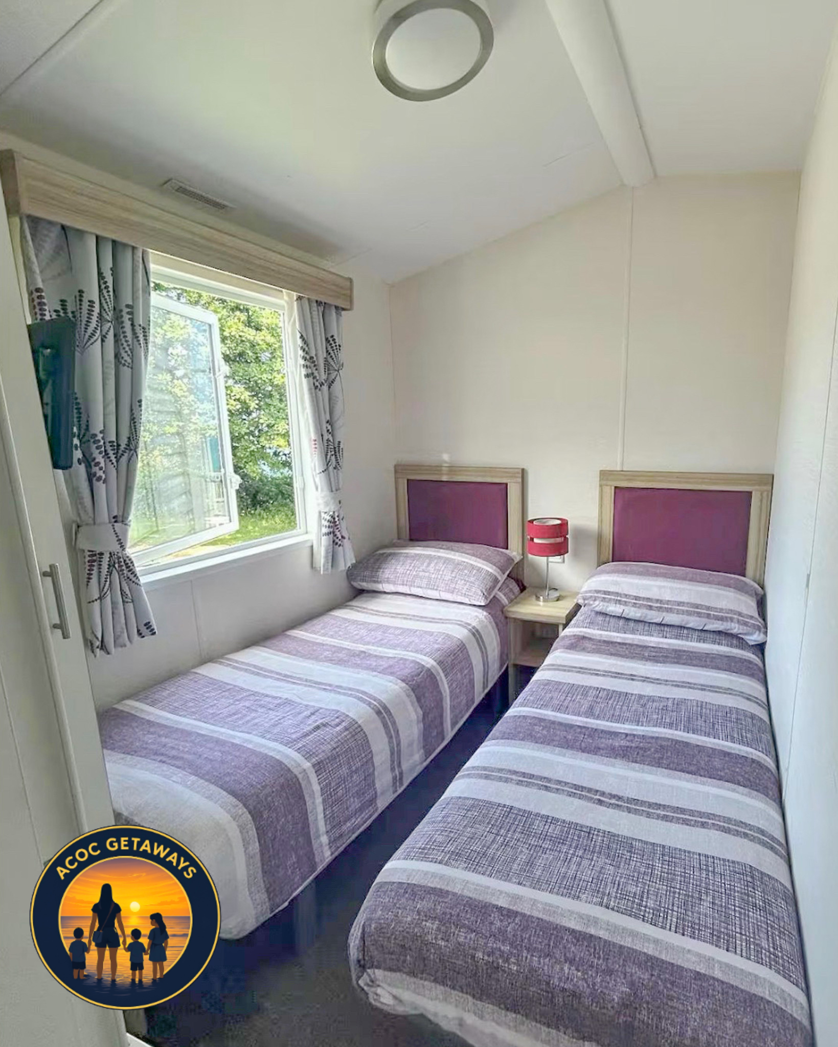 A room with three twin beds with matching plaid bedspreads, a window with curtains showing a green outdoor scene, and a bedside table with a pink lamp.
