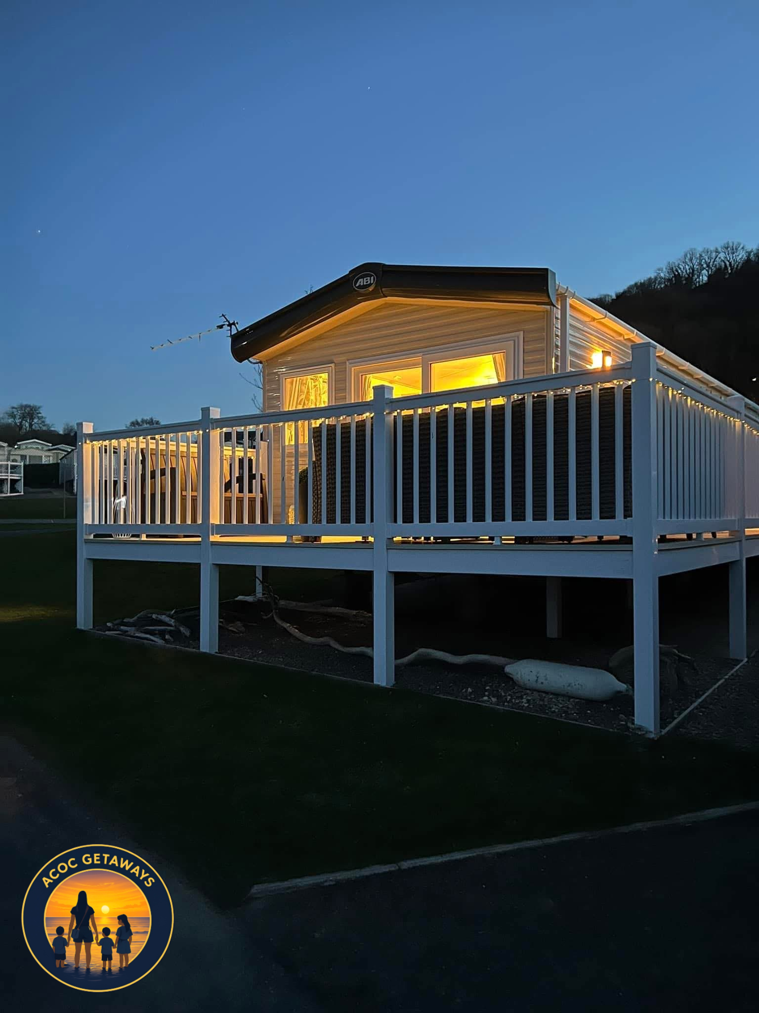 A house at dusk with warm yellow lighting visible through windows, surrounded by a white fence with string lights, and a hillside in the background.
