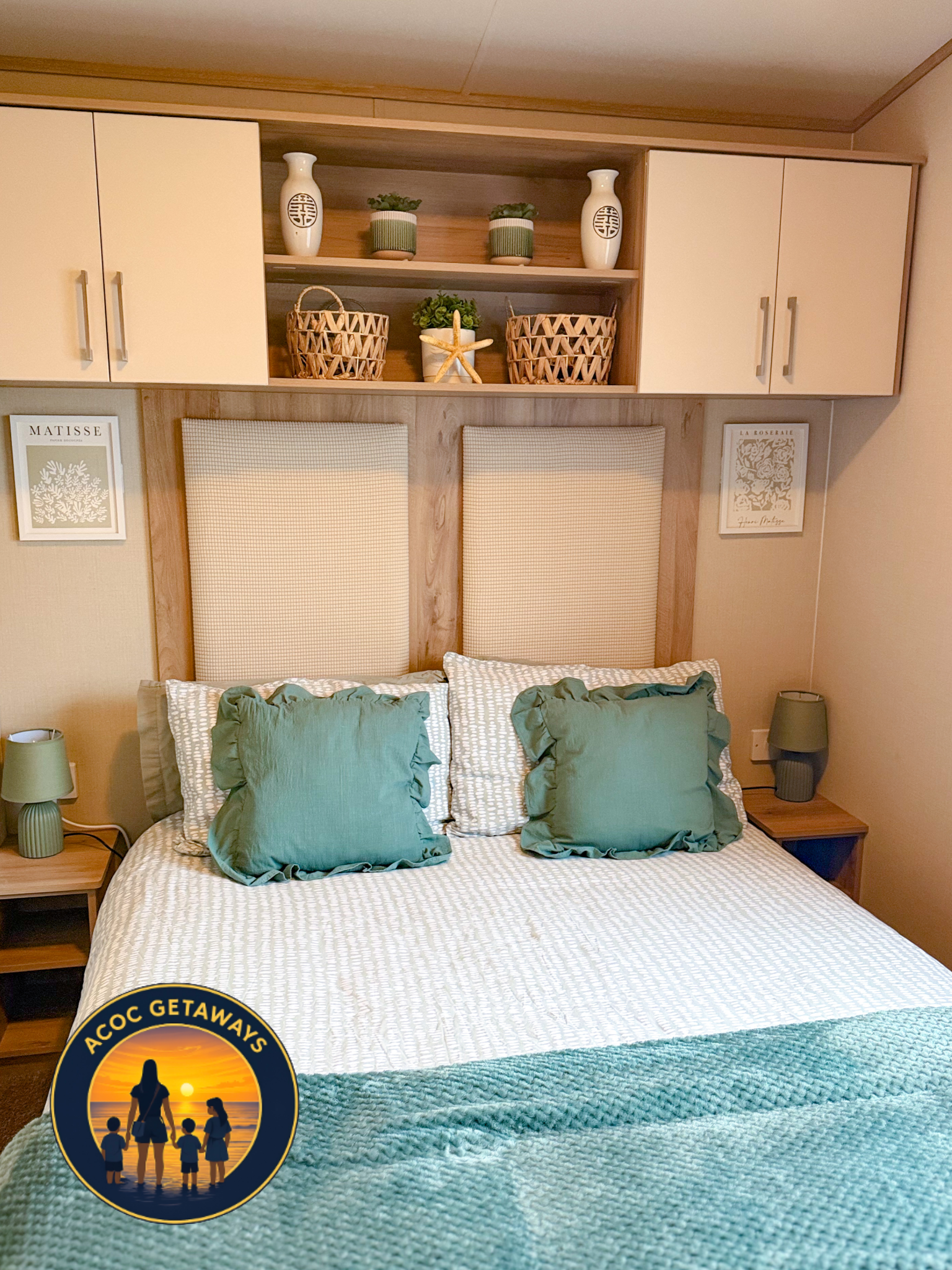 A neatly made bed with four green pillows on a textured white bedspread, two bedside tables with matching green lamps, and a wooden headboard. Above the bed is a shelf with decorative vases, potted plants, wicker baskets, and framed artwork on the wa
