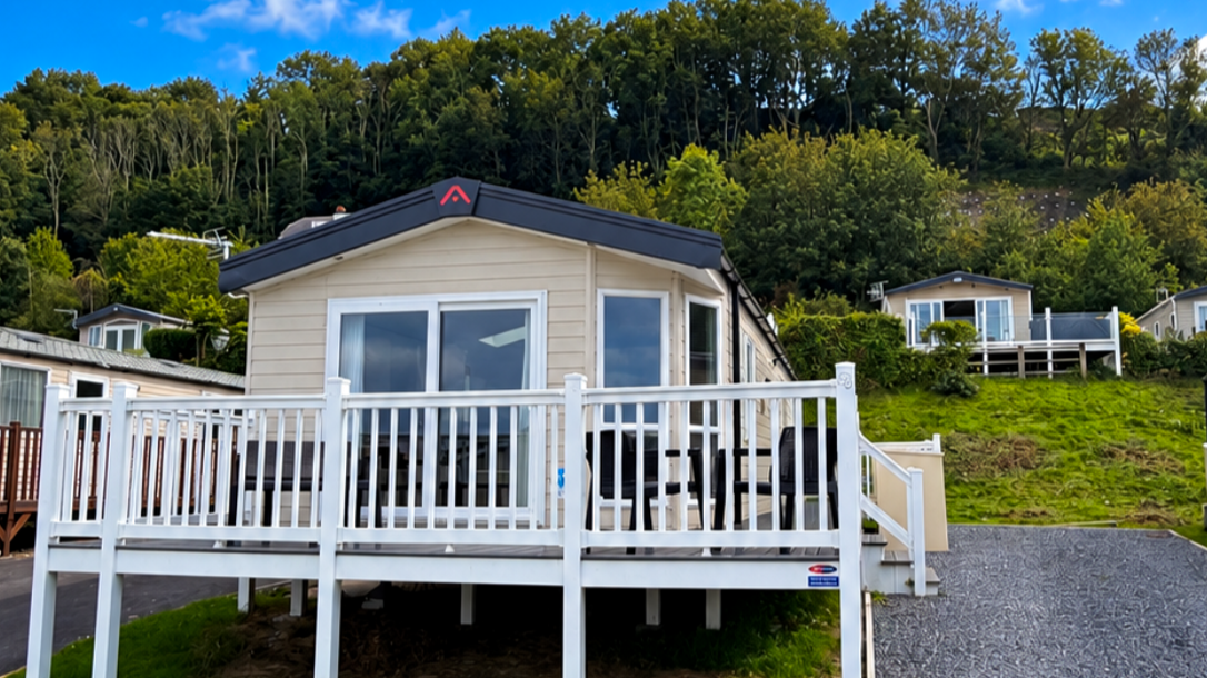 A beige mobile home with large windows and a raised deck with a table and chairs, situated on a grassy area with other similar homes and green trees on a hillside in the background under a blue sky.
