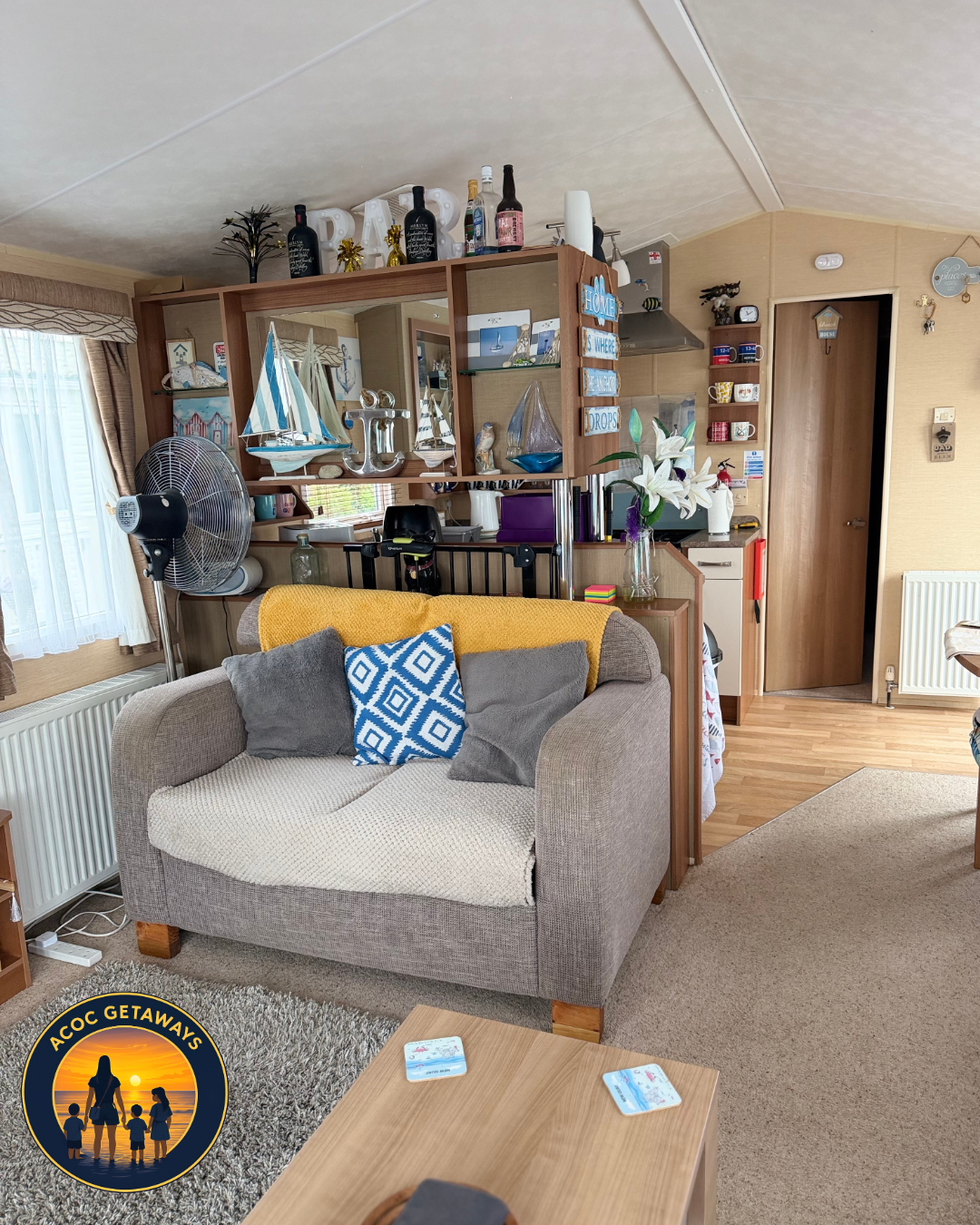 Living room with beige couch, gray and blue pillows, yellow blanket, wooden cabinet with nautical decor, white lilies in glass vase, fan, and miscellaneous household items, doorway, and window with curtains.