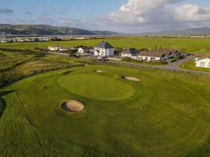 A golf course with sand traps, next to a residential neighborhood with houses and green fields in the background under a partly cloudy sky.