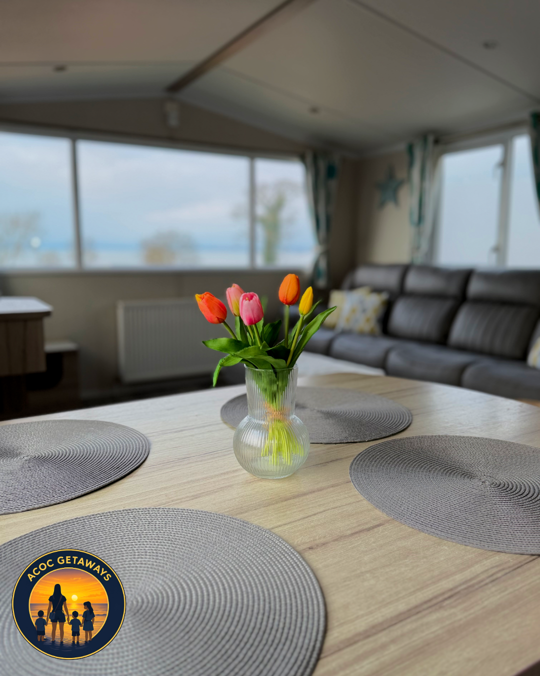 Interior view of a living room with a wooden dining table, a vase of pink, orange, and yellow tulips, gray placemats, a gray sofa with patterned cushions, large windows with curtains, and a scenic outdoor view.