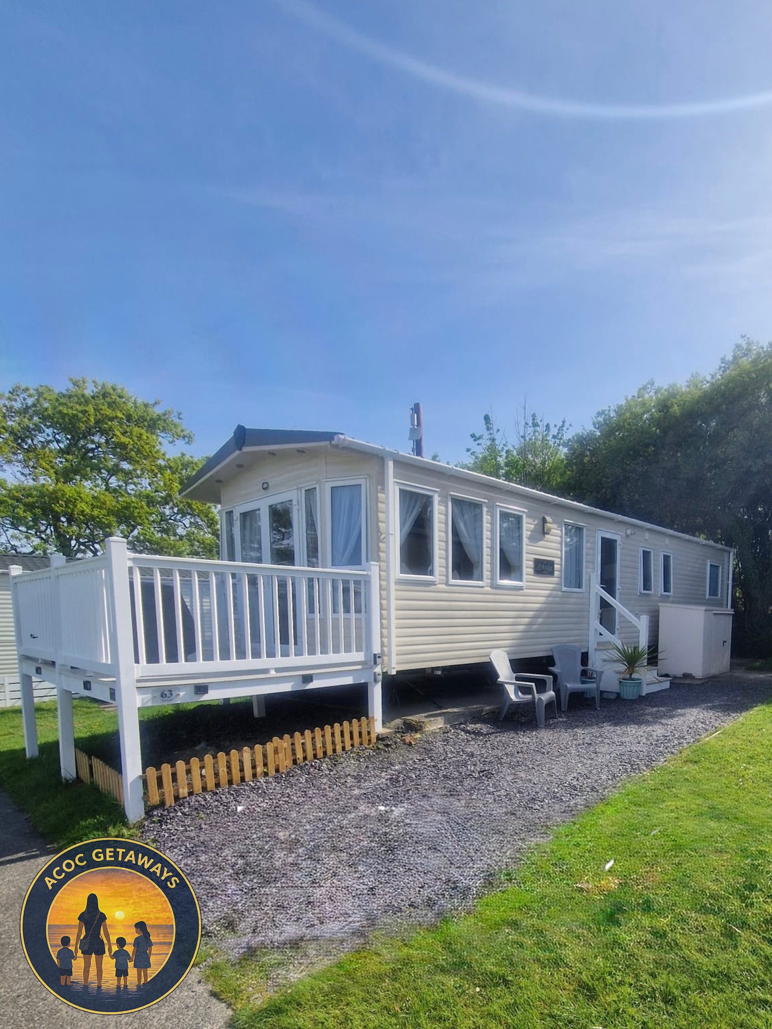 A white modular home with a wooden deck, outdoor chairs, and potted plants, set on a gravel path with grass and trees in the background under a clear blue sky.