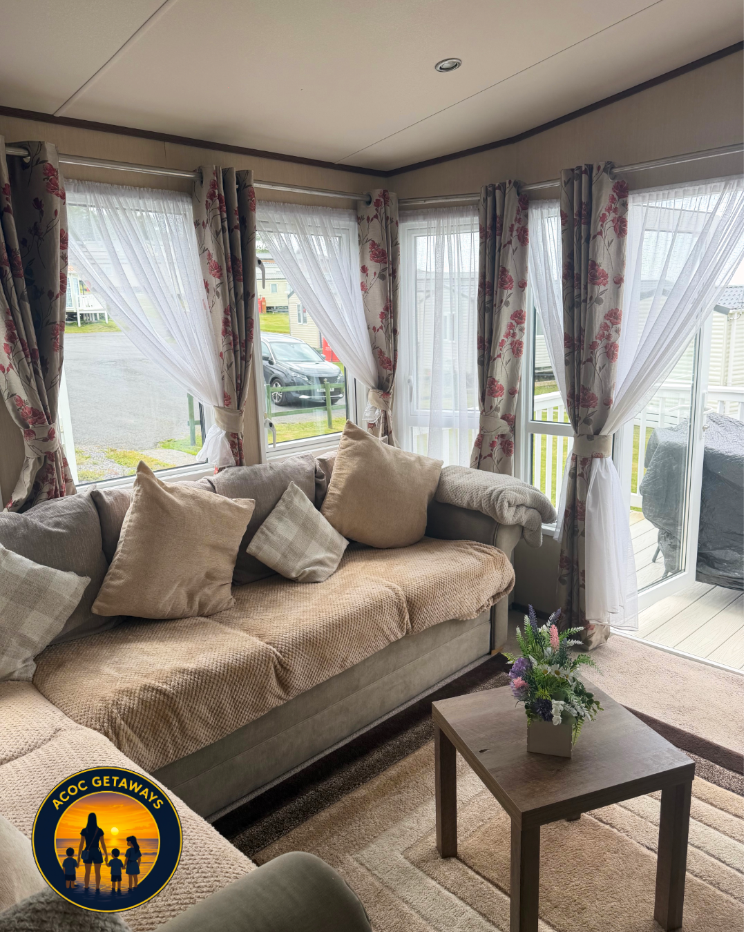 A cozy living room with a beige sofa decorated with multiple cushions. There is a small wooden coffee table with a flower arrangement in a white square vase. The room features large windows with floral curtains and sheer white drapes, allowing natura