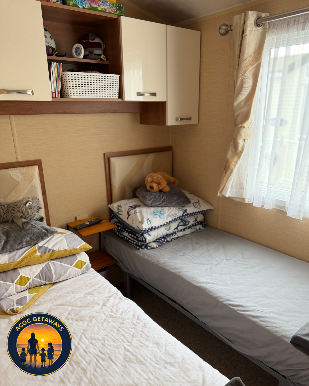 A bedroom with two beds, one with stuffed animals, plush toys, and patterned bedding, and a window with beige curtains allowing natural light.