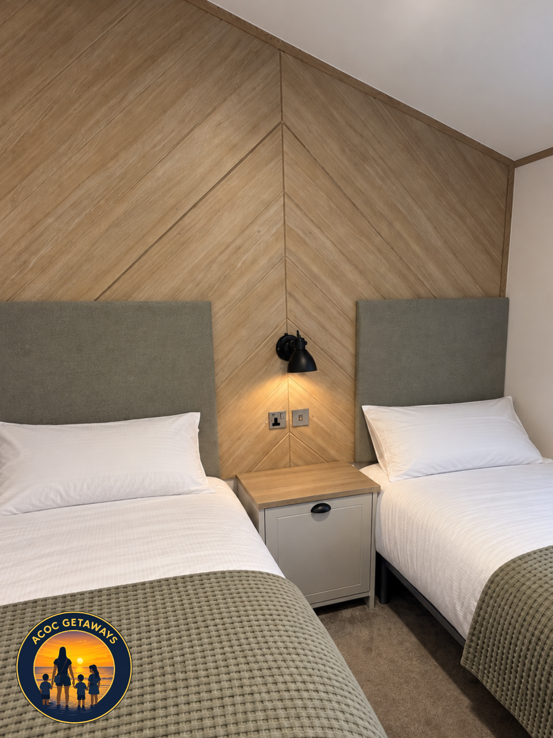 Two beds with gray headboards and white linens in a hotel room, separated by a nightstand with a black wall lamp attached to a wood-paneled wall.