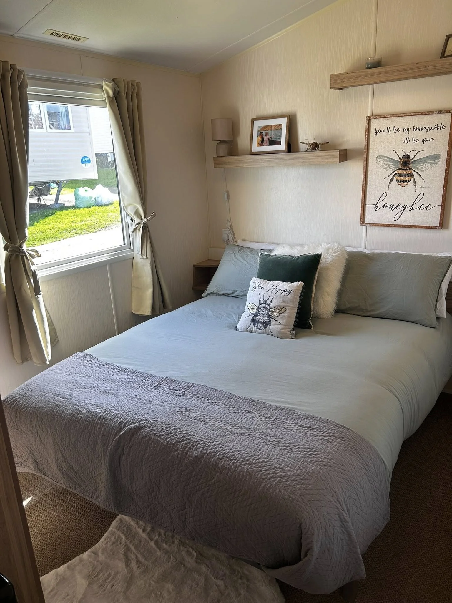 A cozy bedroom with a large bed that has gray bedding and decorative pillows featuring a bee and the words "Bee Happy". There's a window with beige curtains drawn to the sides, revealing a grassy outdoor area and a white trailer outside. Above the be