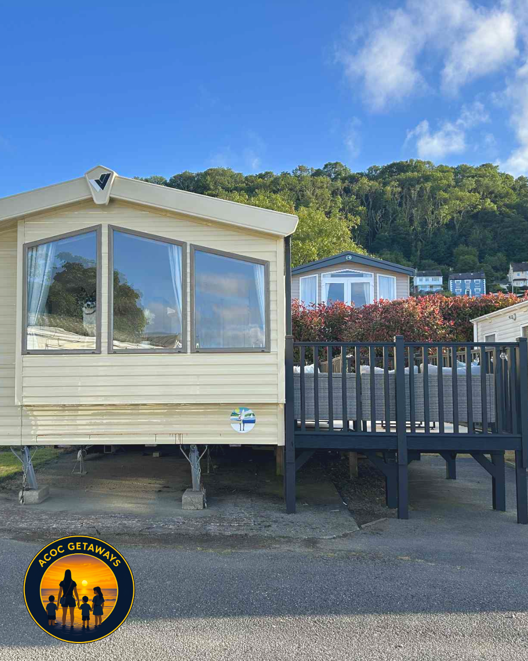 A small beige mobile home with large front windows reflecting trees and sky, elevated on blocks with a black wooden deck and stairs, surrounded by a residential neighborhood with lush green hills in the background, under a bright blue sky with some c