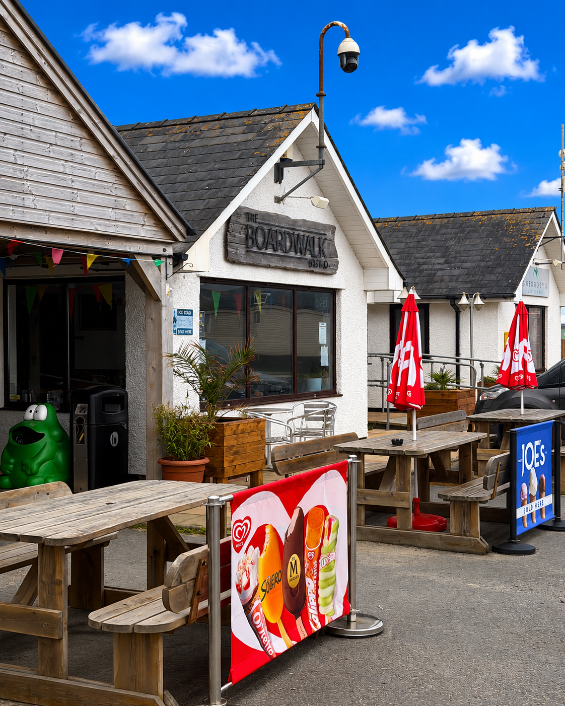 Exterior of The Boardwalk Bistro with outdoor seating, red umbrellas, advertising banners for ice cream, a green frog-shaped trash bin, and a parking lot under a blue sky with scattered clouds.