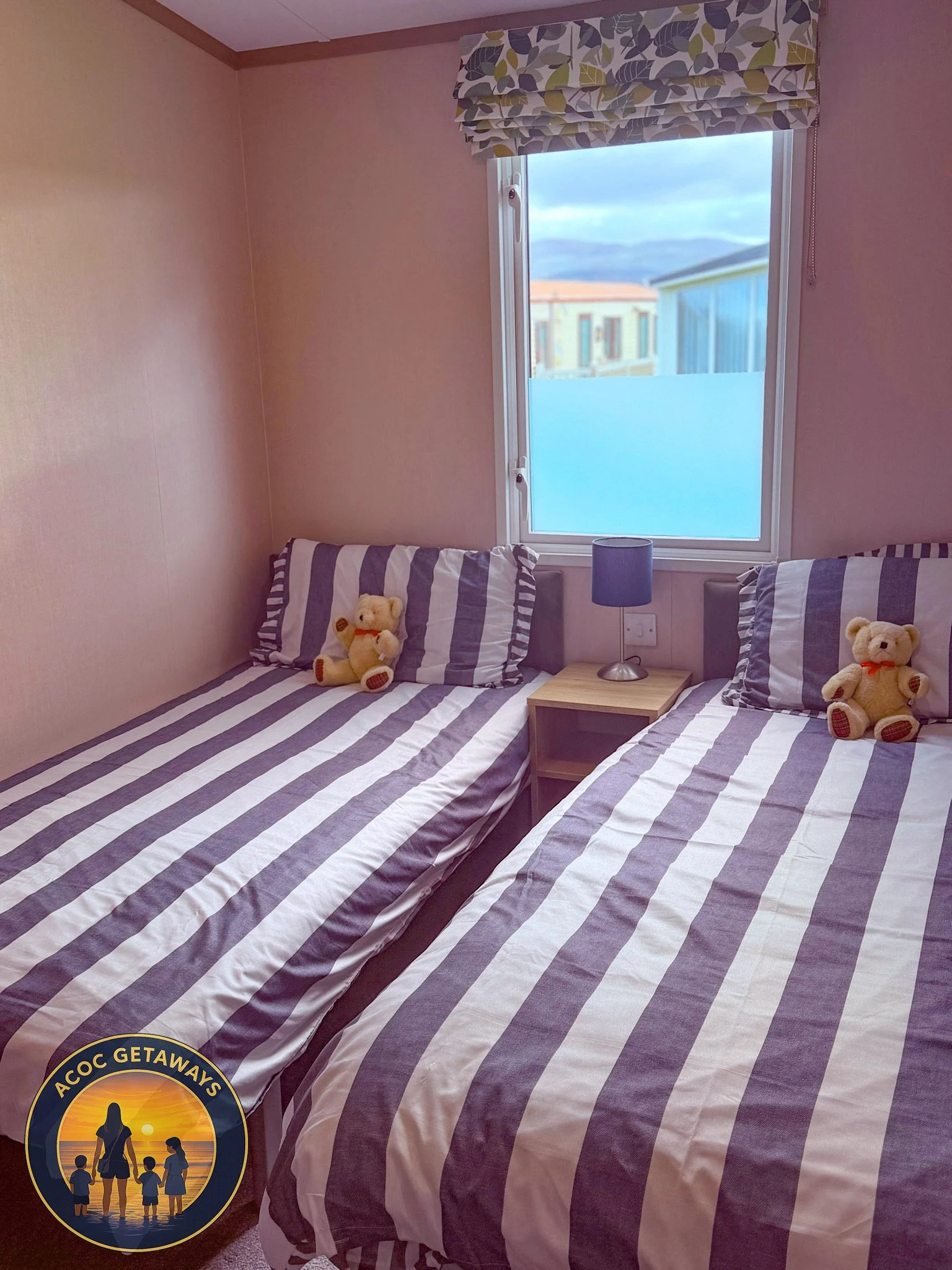 Two twin beds with striped bedspreads and pillows, each with a teddy bear, positioned beside a window with a patterned valance, and a small wooden nightstand with a blue lamp between the beds.