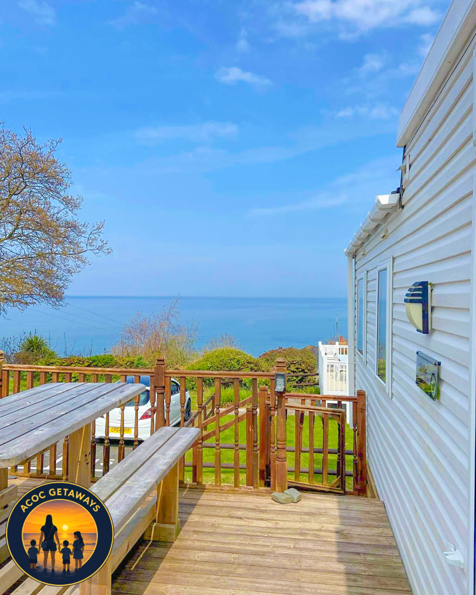View of a wooden deck, part of a house with white siding, overlooking a landscape with bushes, trees, and the ocean under a partly cloudy sky.