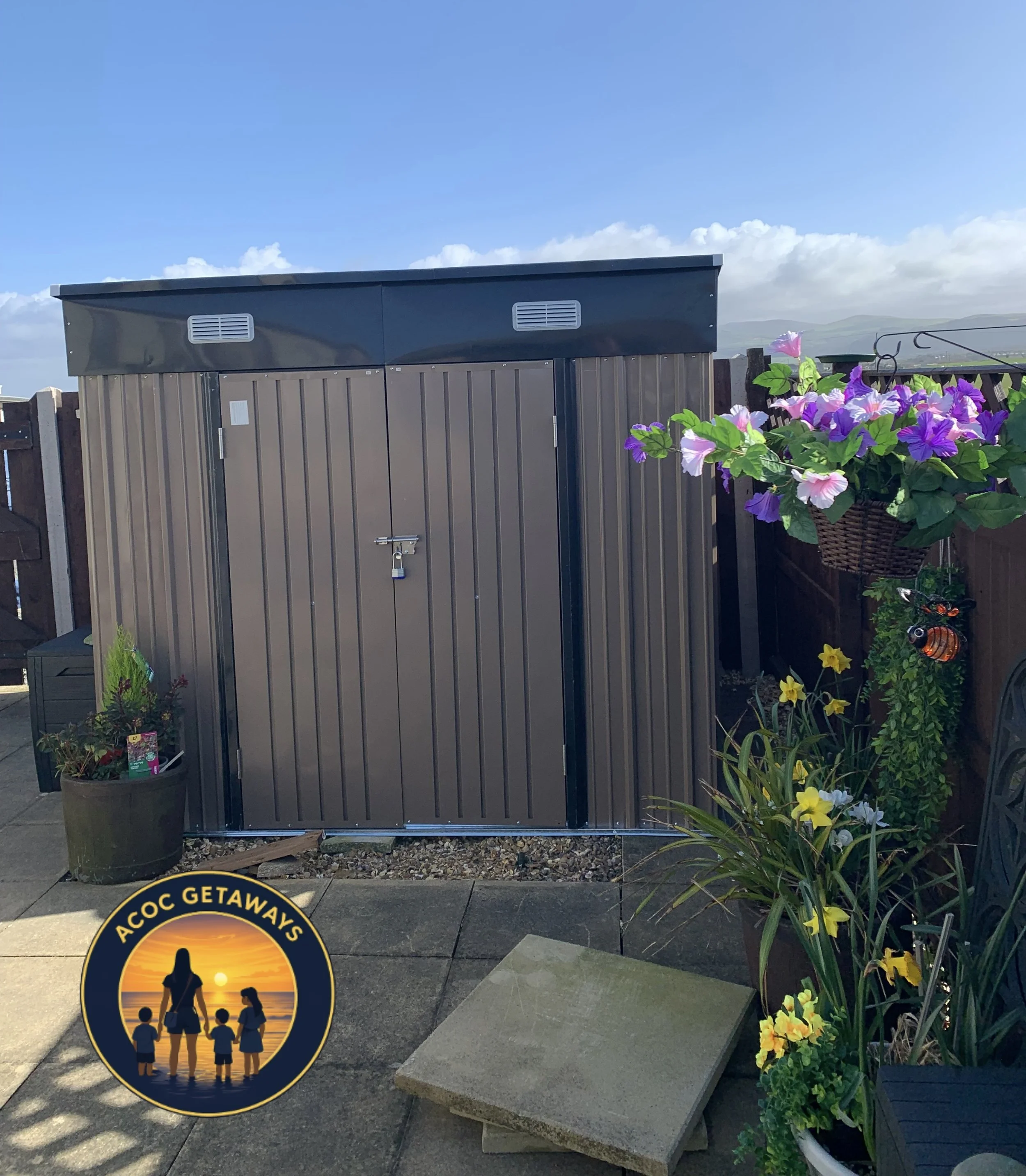 A brown metal outdoor shed with double doors, situated on a paved patio area. To the right, there are vibrant purple and white flowers in a hanging basket, and to the left, a large potted plant. There is an ACoc Getaways logo featuring a family silho