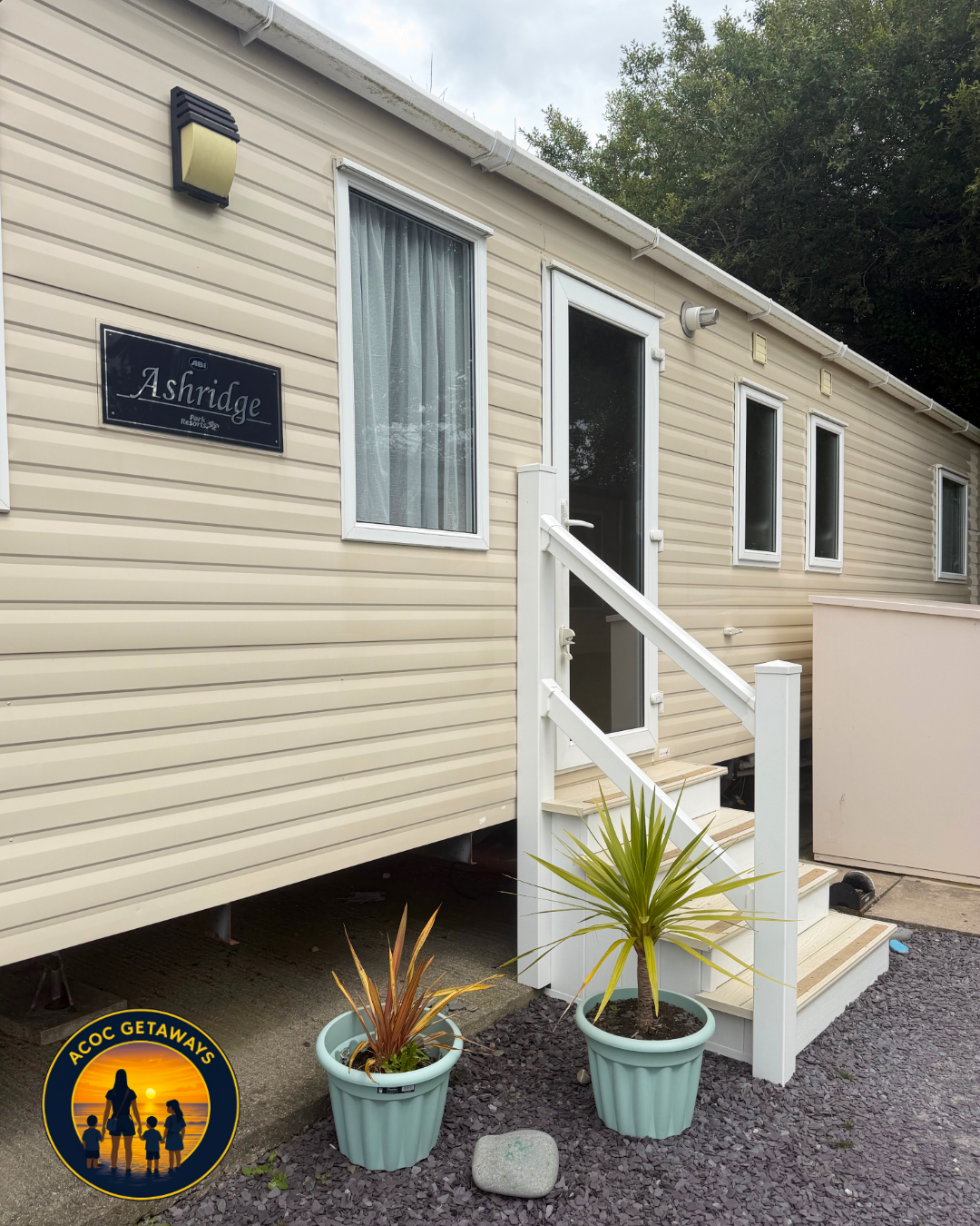 Exterior view of a beige mobile home with three windows and a door, labeled Ashridge. There are two potted plants at the foot of the stairs leading to the door, and a logo in the bottom left corner features a sunset scene with a woman and children, l