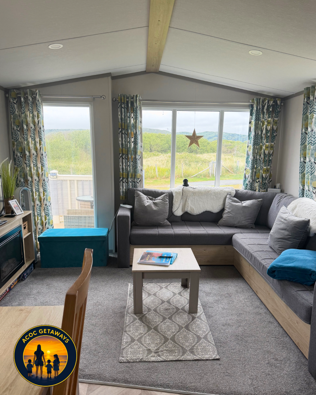 Living room with large windows showing a grassy landscape and mountains in the background. There is a dark gray L-shaped sectional sofa with gray and white pillows, a white blanket, and a blue folded blanket. A small white coffee table with magazines