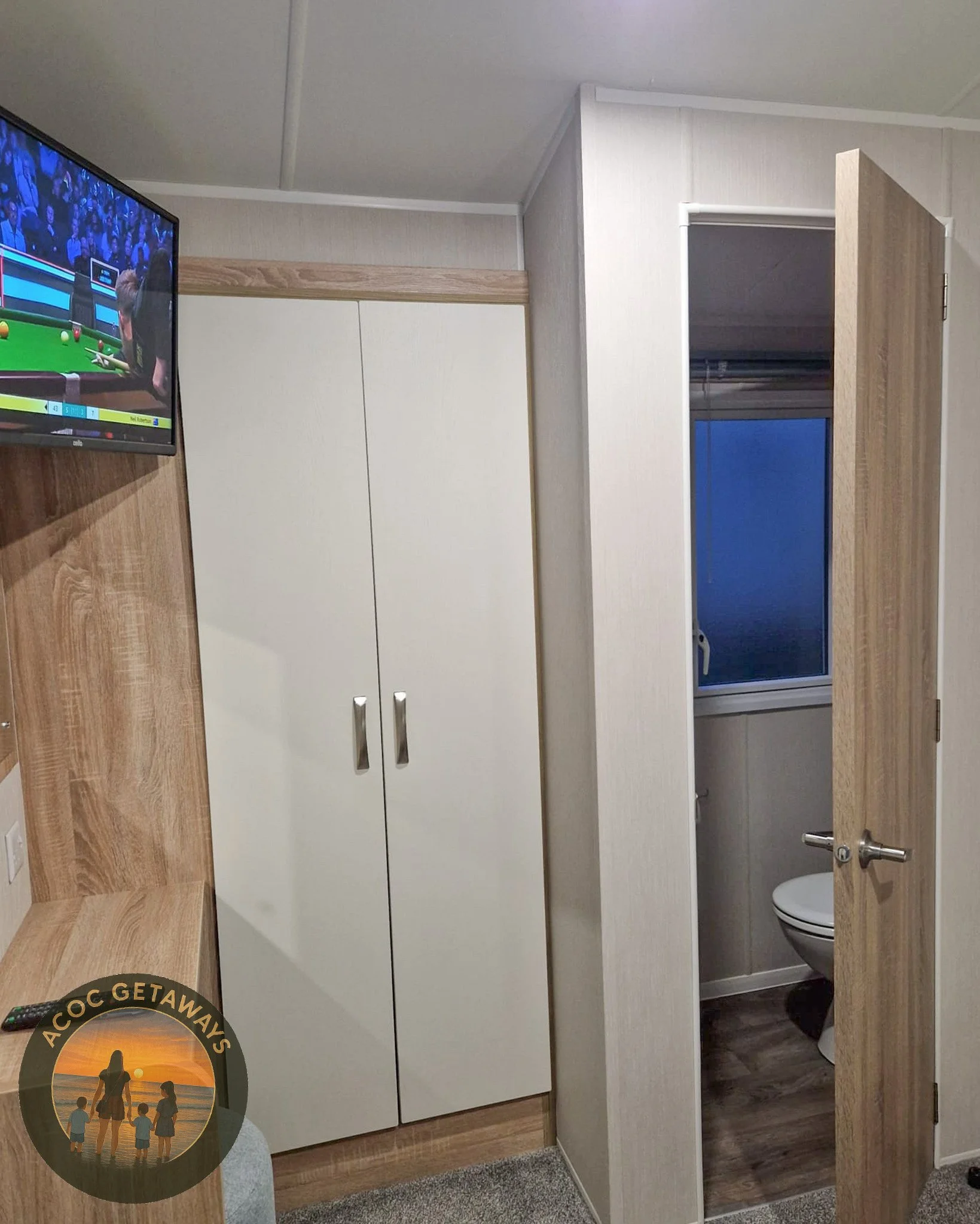 A room with a mounted TV showing a sporting event, a white closet with metal handles, a wooden desk or shelf, and a door slightly open revealing a bathroom with a toilet and a window.