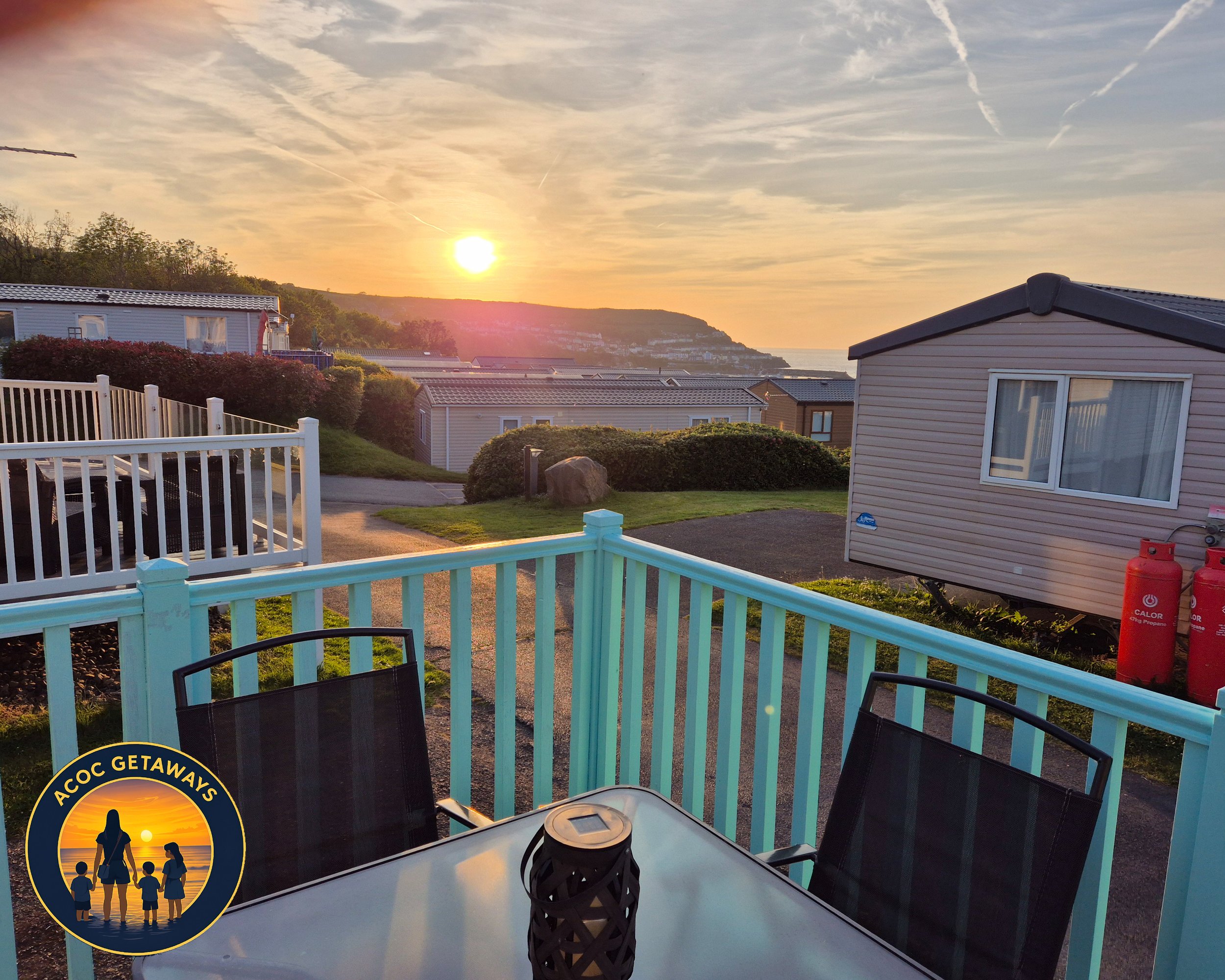 Sunset view from a balcony with black chairs and a table, overlooking a coastal neighborhood with mobile homes and a hill in the distance.