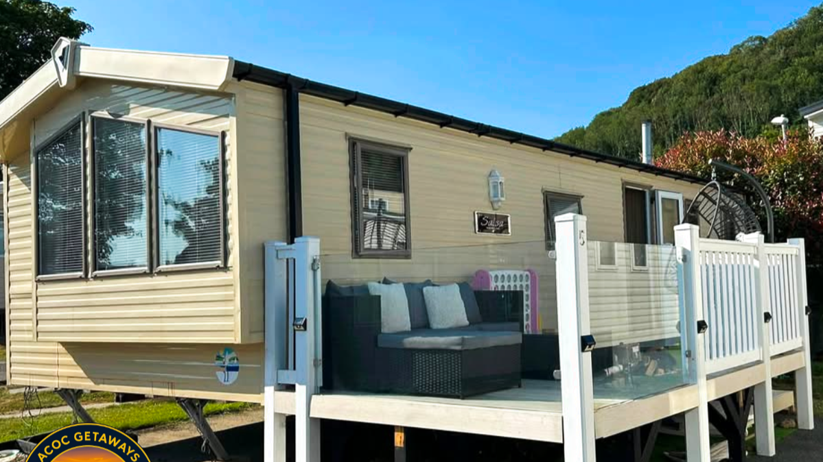 A beige mobile home with a small elevated porch that has white railings and glass panels. The porch has a black outdoor sofa with light-colored pillows. There is a sign on the wall that says "Sailor," and a light fixture above it. Behind the mobile home, there is a green hillside under a blue sky.