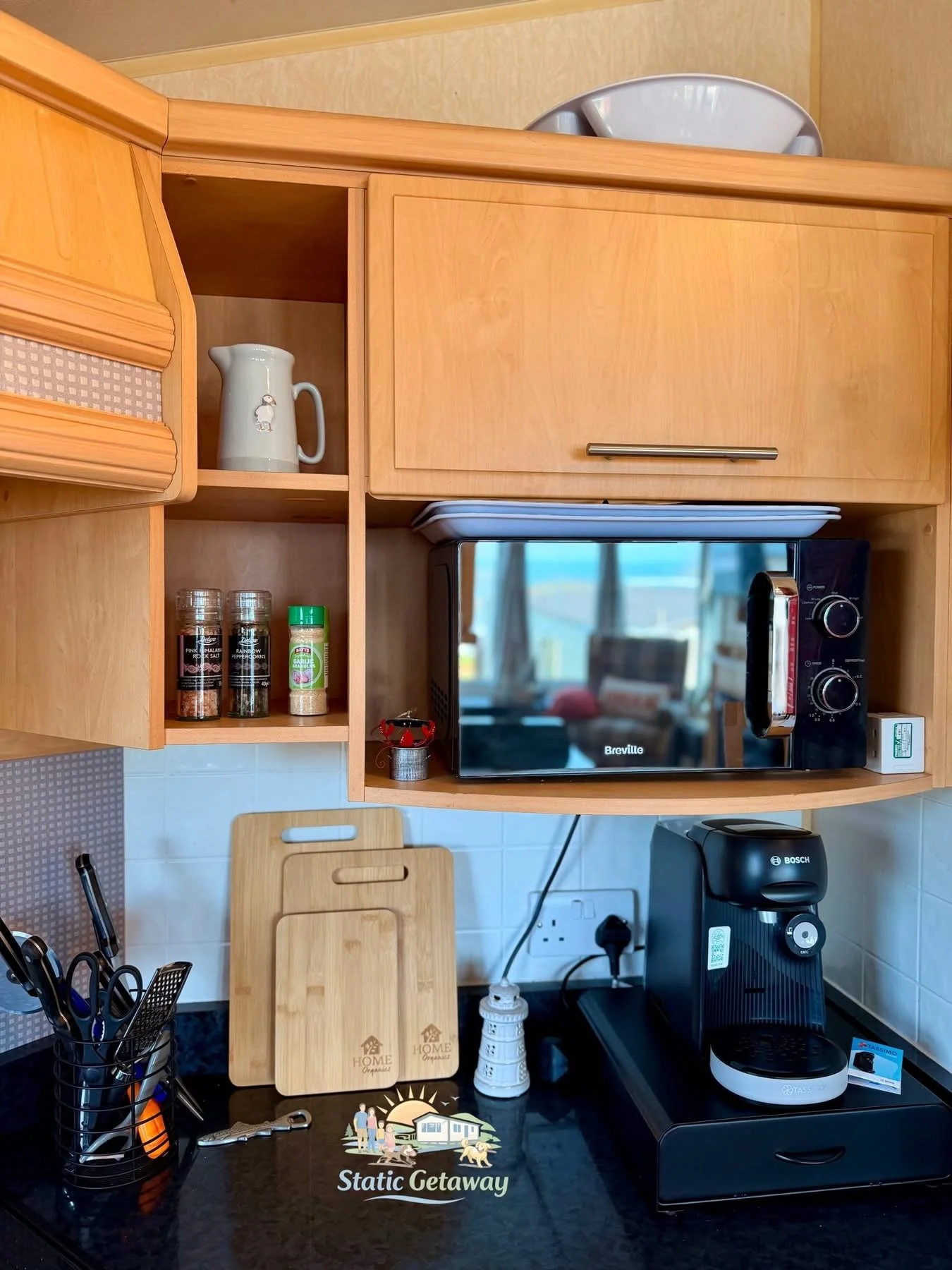 A kitchen countertop with a black coffee machine, a set of wooden cutting boards, and a container with various knives. Above, wooden kitchen cabinets with a microwave oven inside one, and spice jars on an open shelf.