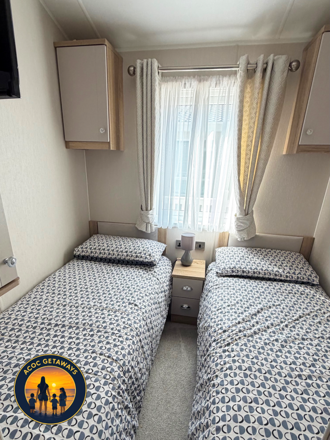 A small bedroom with two twin beds with matching patterned bedspreads, separated by a nightstand with a lamp, a window with white sheer curtains and decorative drapes, and mounted cabinets above the beds.