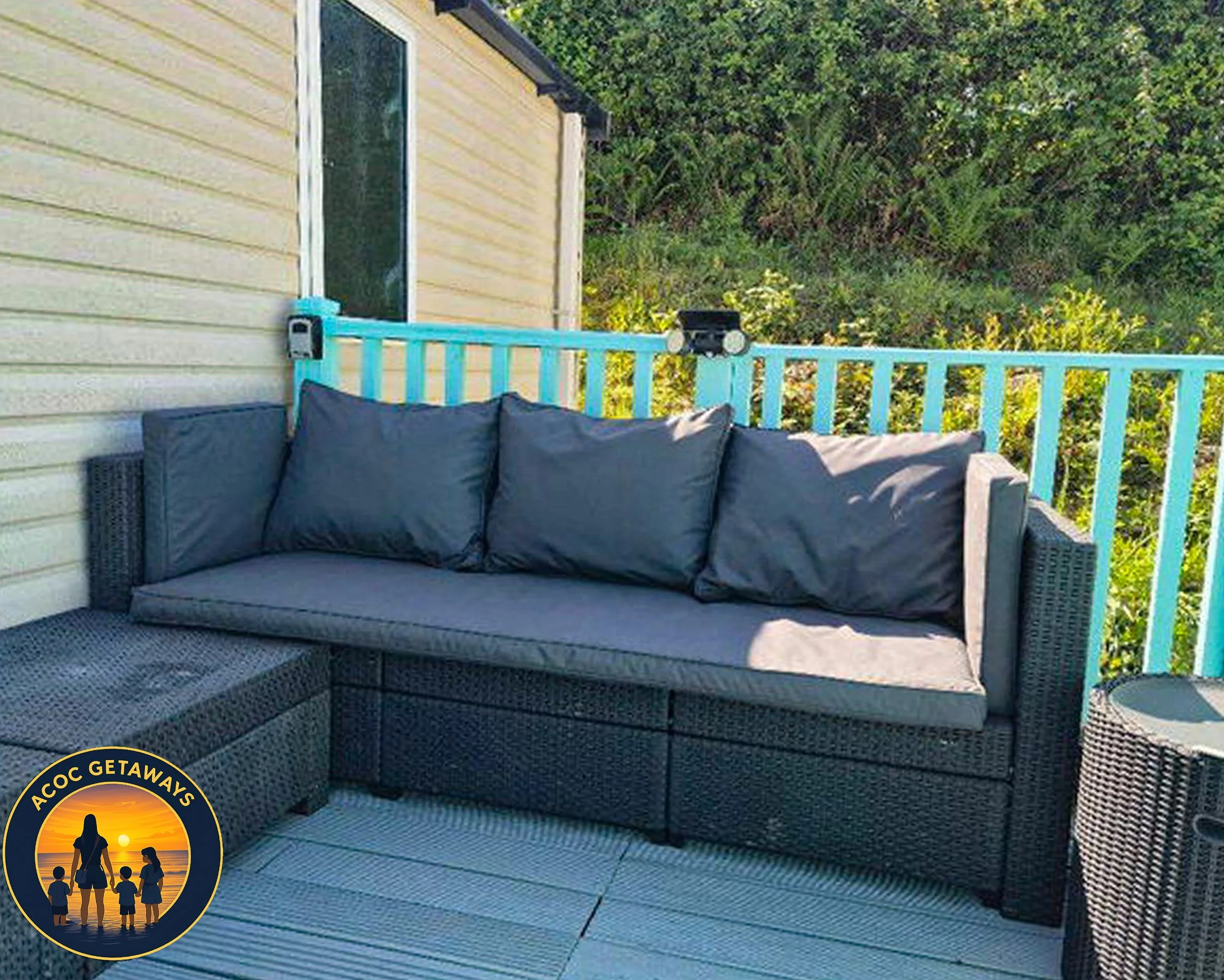 Outdoor patio area with a black wicker sofa with four dark gray cushions, a matching wicker side table, a beige house with vinyl siding, a blue railing, and green trees in the background.
