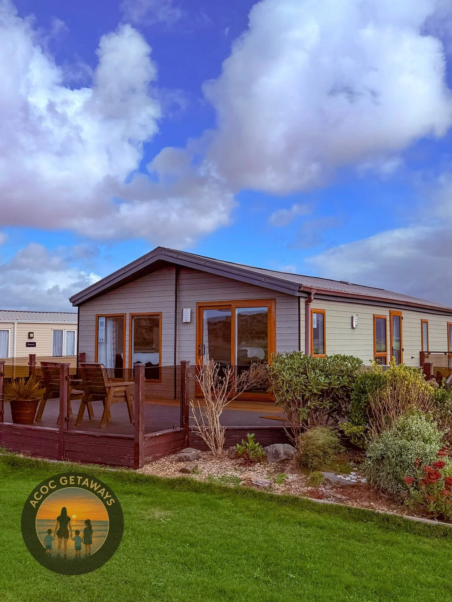 A small cottage-style home with a wooden deck and garden, under a partly cloudy sky. There is a lawn in front with some bushes and a logo in the lower left corner reading "ACOC GETAWAYS" with an image of a woman and children at the beach.