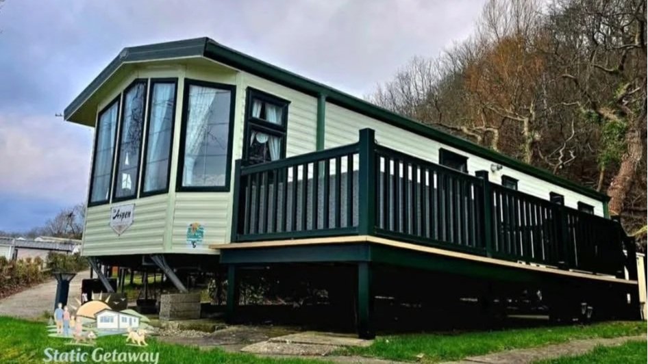 A small elevated house with beige siding, black window frames, and a black railing on the deck, situated on a grassy area with trees in the background.