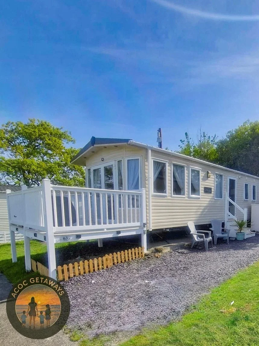 Vacation rental mobile home with a deck, outdoor chairs, and a small fence, on a sunny day with clear blue sky and green trees.