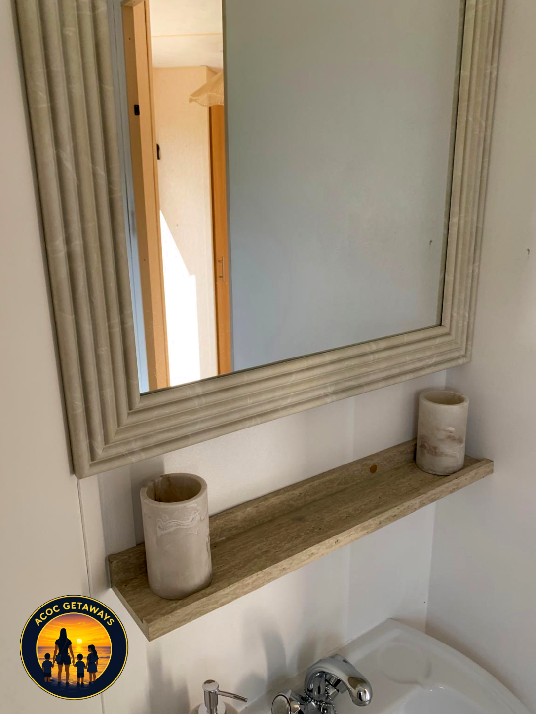 A bathroom mirror with a carved wooden frame, a wooden shelf below it holding two ceramic candle holders, and part of a bathtub with a sink faucet visible at the bottom.