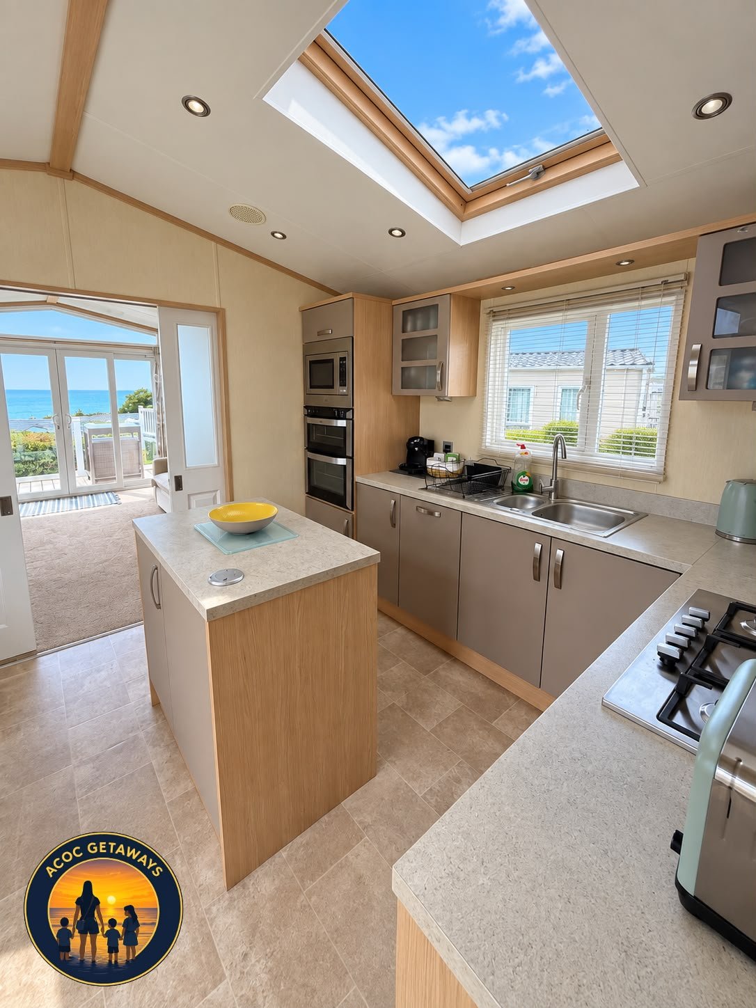 Kitchen with skylight, window, and view of outdoor patio and ocean, featuring beige and gray cabinets, microwave, oven, coffee maker, sink, stovetop, and countertop with a yellow bowl.