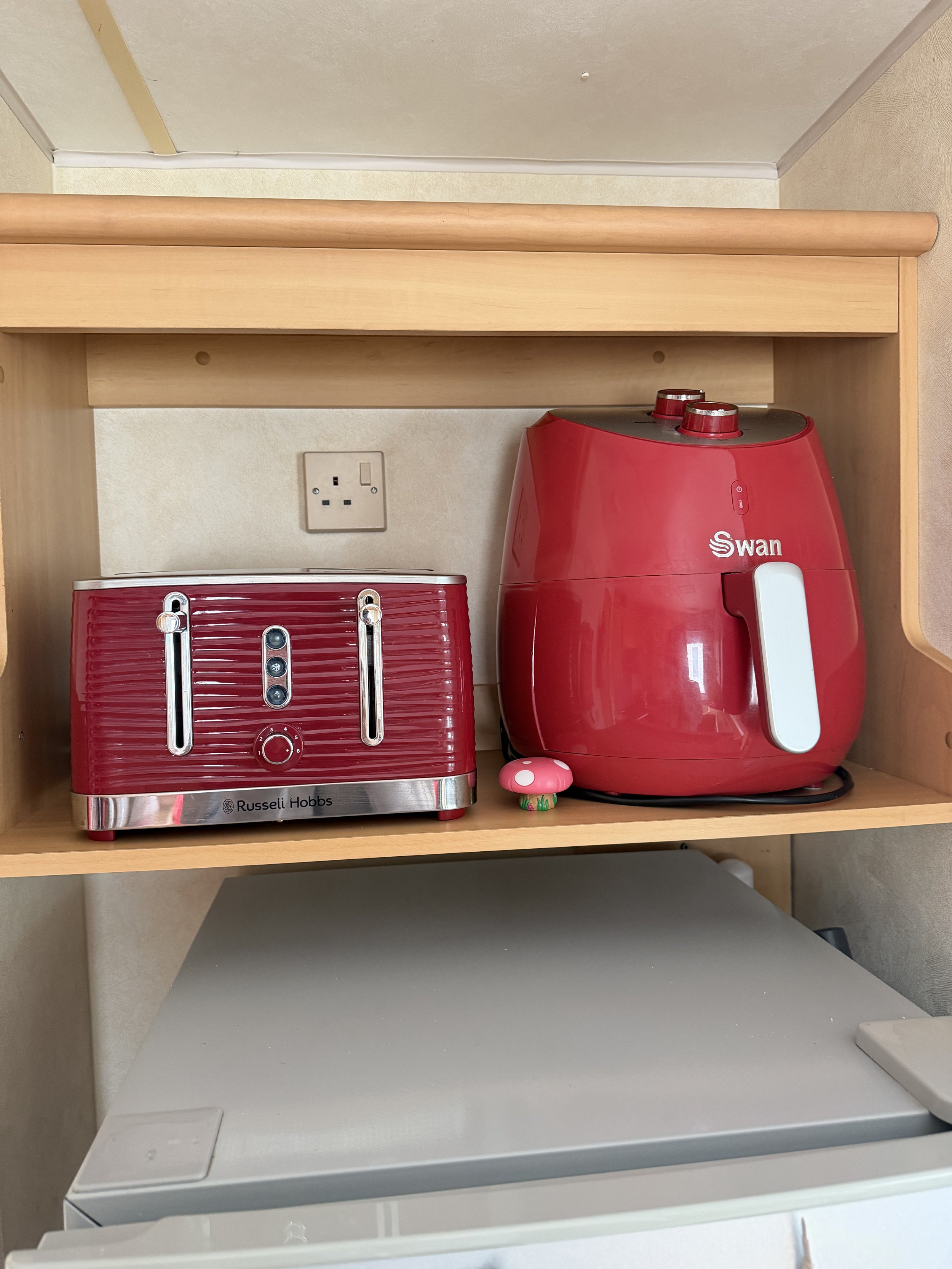 A red Russell Hobbs toaster oven and a red Swan air fryer on a wooden shelf with an electrical outlet in the background.