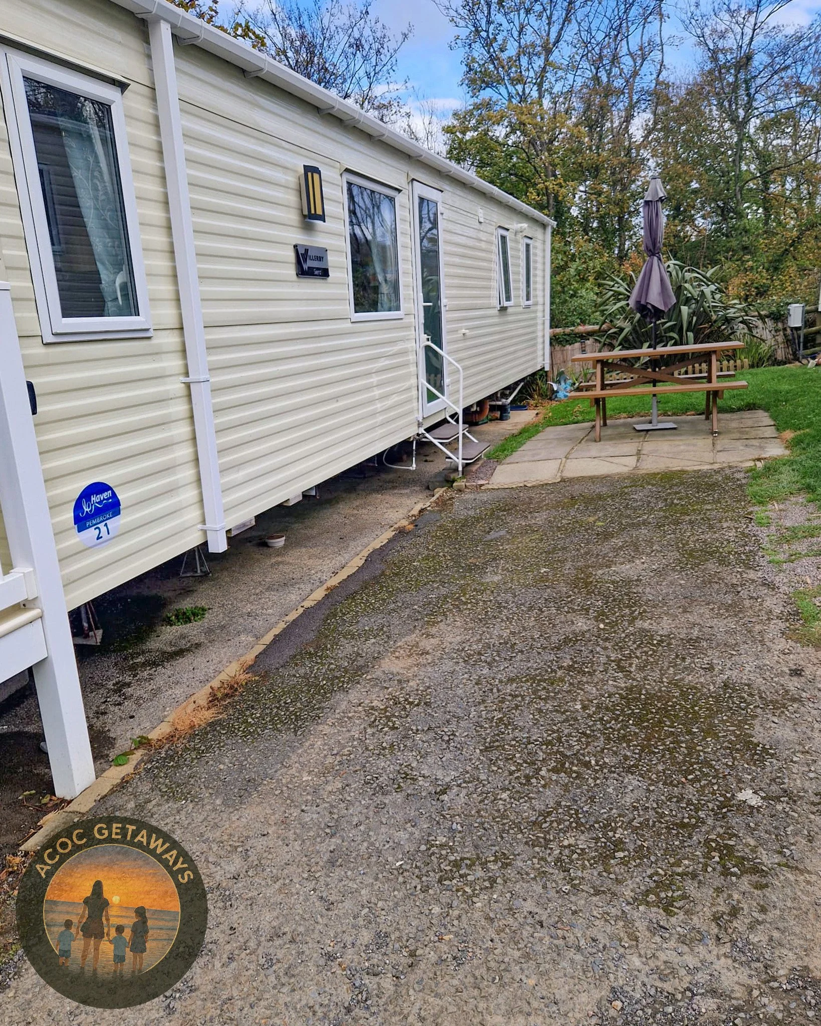 A beige trailer with white trim, three windows, and a small entrance with a white handrail. There is a small patio area with a picnic table, to the right, with a closed gray umbrella. The ground in front of the trailer is a mix of gravel and concrete