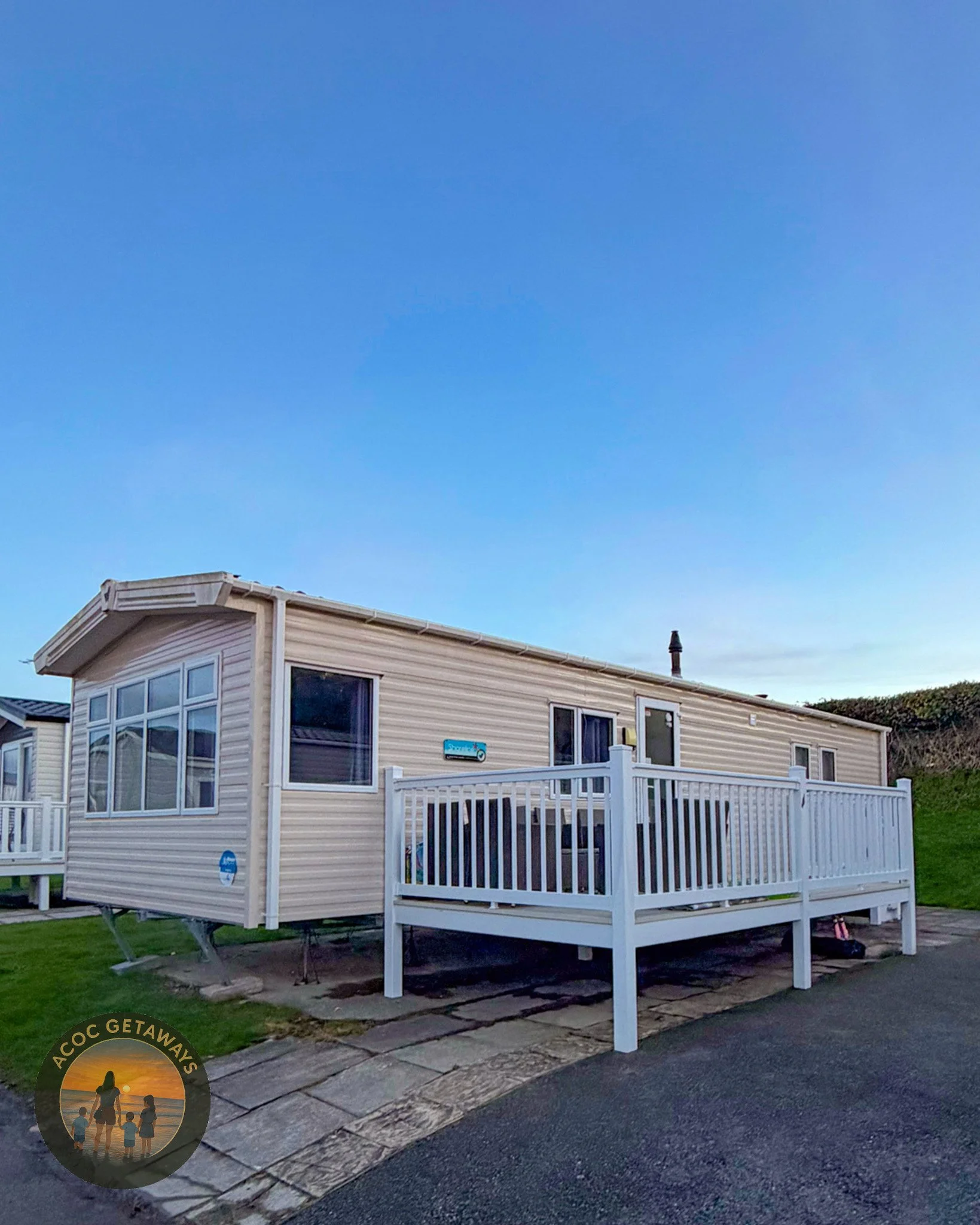 A beige mobile home with large front windows and a white balcony railing, situated on a paved area with a grassy lawn nearby, under a clear blue sky.