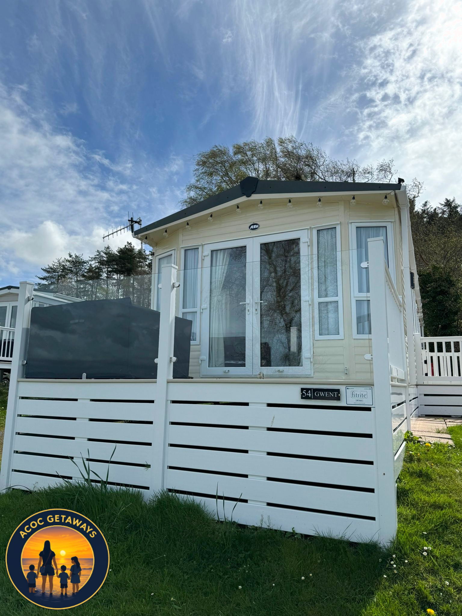 A small, white, prefabricated house with glass doors and windows, situated on a grassy lawn with a white fence in front, under a partly cloudy sky with trees in the background.