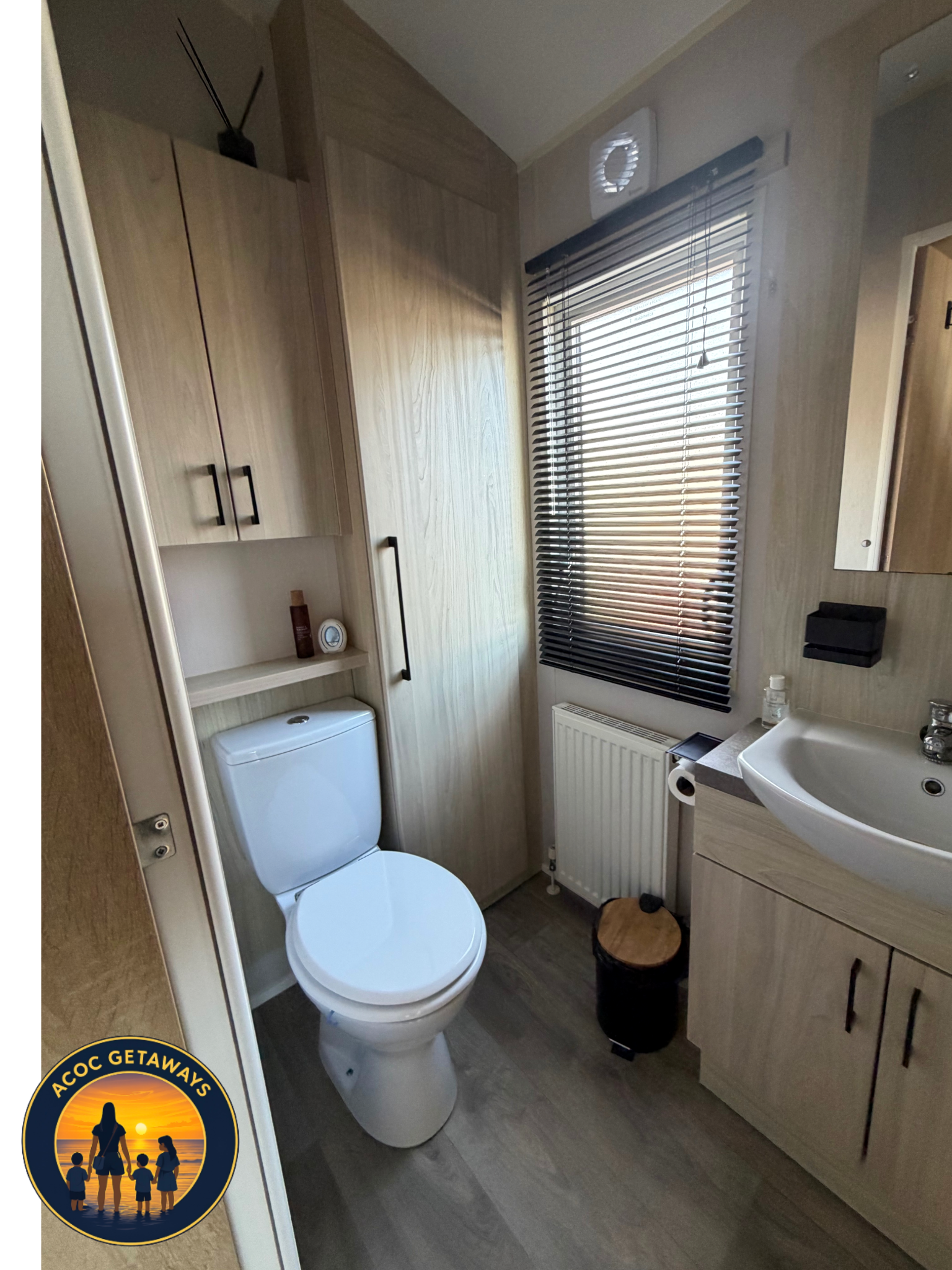 A small bathroom featuring a toilet, a sink with a cabinet below, a wooden storage unit with black handles, a window with blinds, a heater, a small black container, a bottle of hand sanitizer, a wastebasket with a wooden lid, and a black wall-mounted