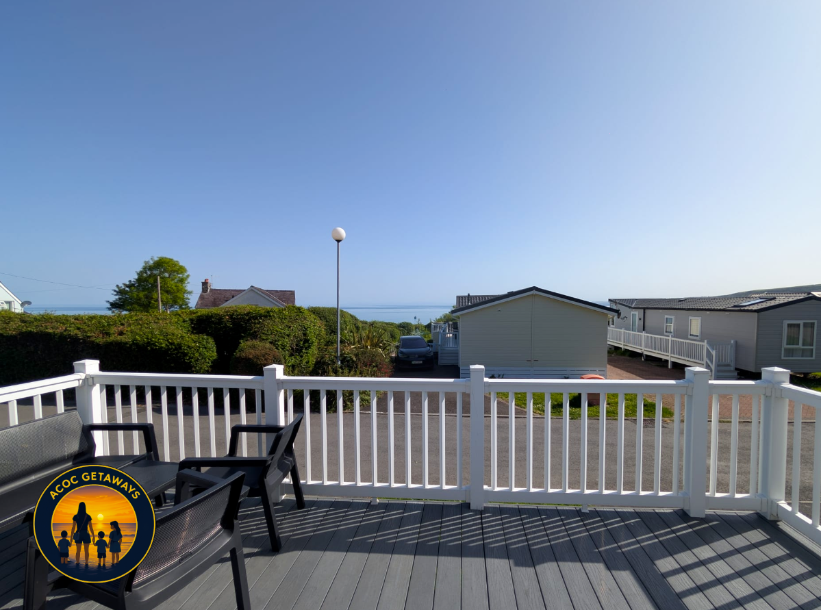View from a deck with outdoor furniture, overlooking a small residential area with mobile homes and bushes, with a clear blue sky and the ocean in the distance.