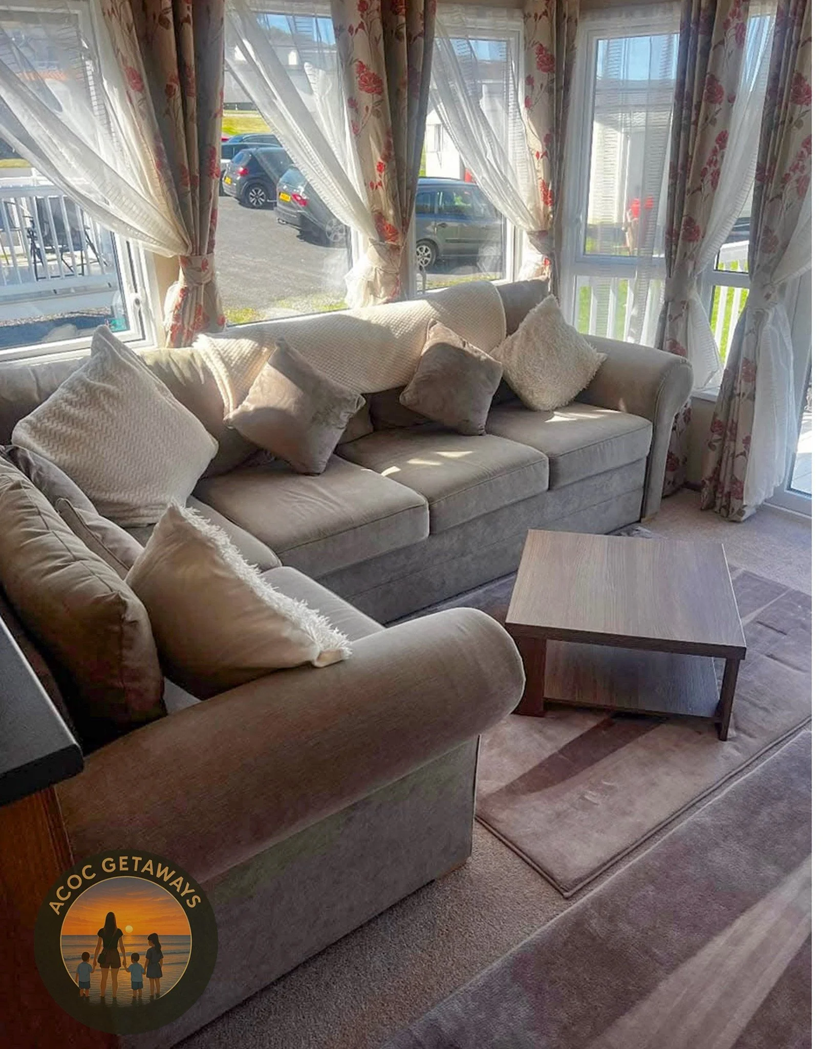 Living room with two beige sofas, multiple pillows, a wooden coffee table, and large windows with floral curtains letting in sunlight.