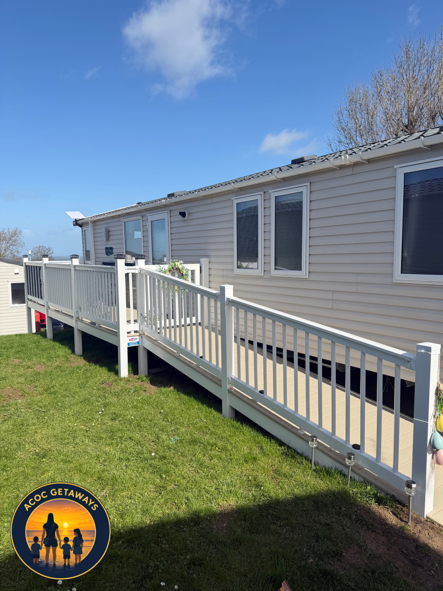A beige mobile home with four windows and a small deck with a white railing. There is a flowerpot with plants on the deck and solar lights along the ground next to the deck. The sky is blue with a few clouds, and there are leafless trees in the backg