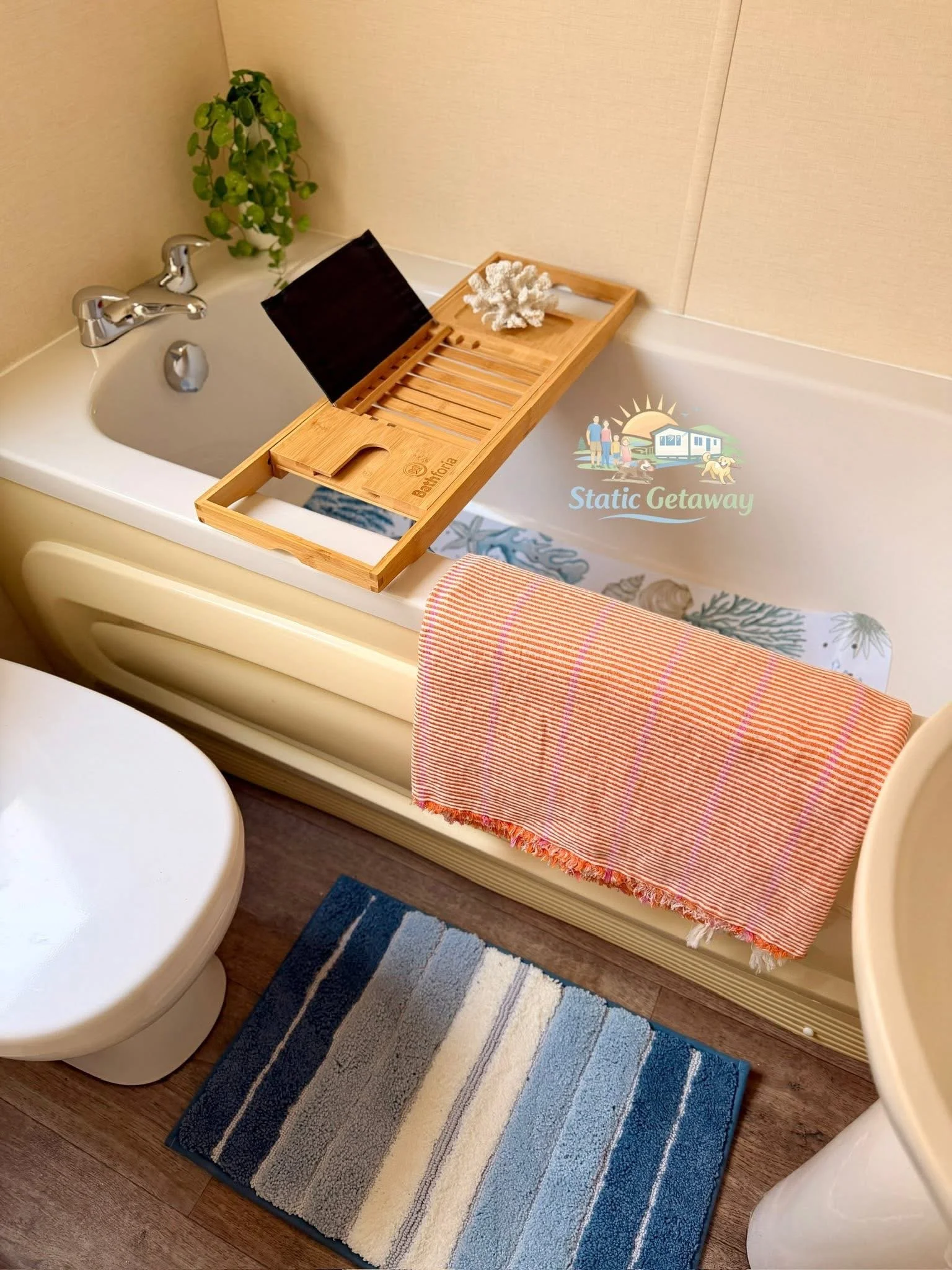 A bathroom scene showing a small rectangular blue, gray, black, and white striped rug on a wood floor, a toilet with closed lid, and a bathtub with a striped orange and white towel hanging over the edge. There is a wooden bath tray holding a white co