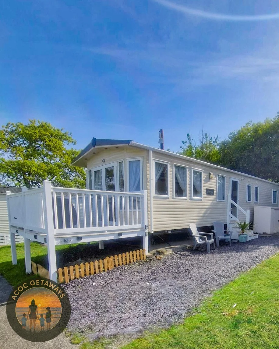 A beige mobile home with a white raised porch and stairs, outdoor chairs, and a potted plant. The ground is gravel, and there is a green lawn with trees in the background. A logo of a woman and children at sunset with the text "AOC GETAWAYS" is in the bottom left corner.