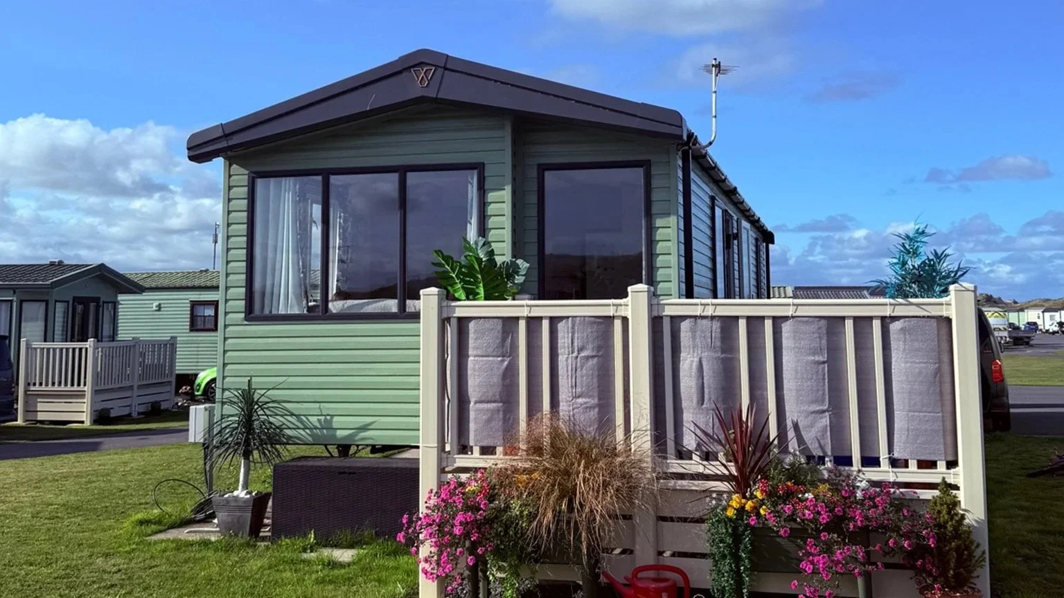A green manufactured home with large window, surrounded by a small yard with plants, flowers, and a white privacy fence, under a partly cloudy blue sky.