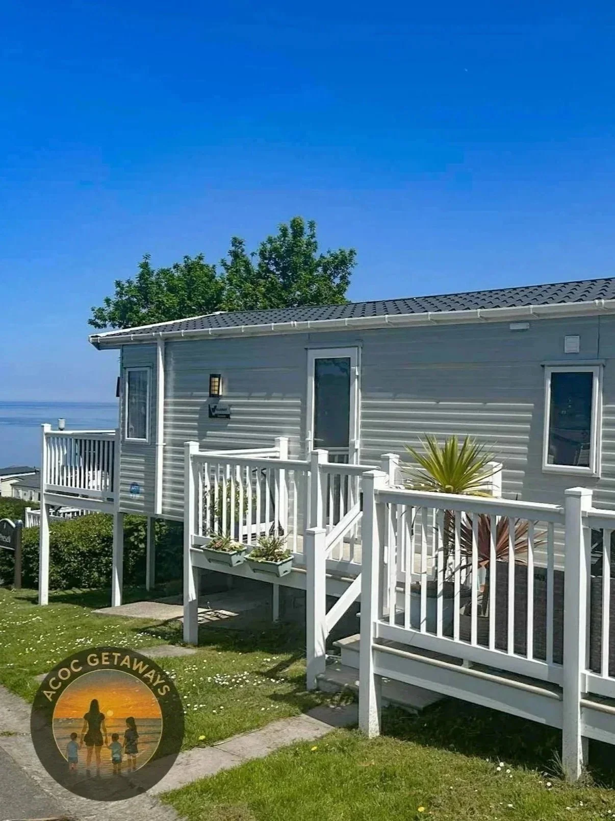A gray house with a white railing porch, green grass, potted plants, and a blue sky with a few clouds.